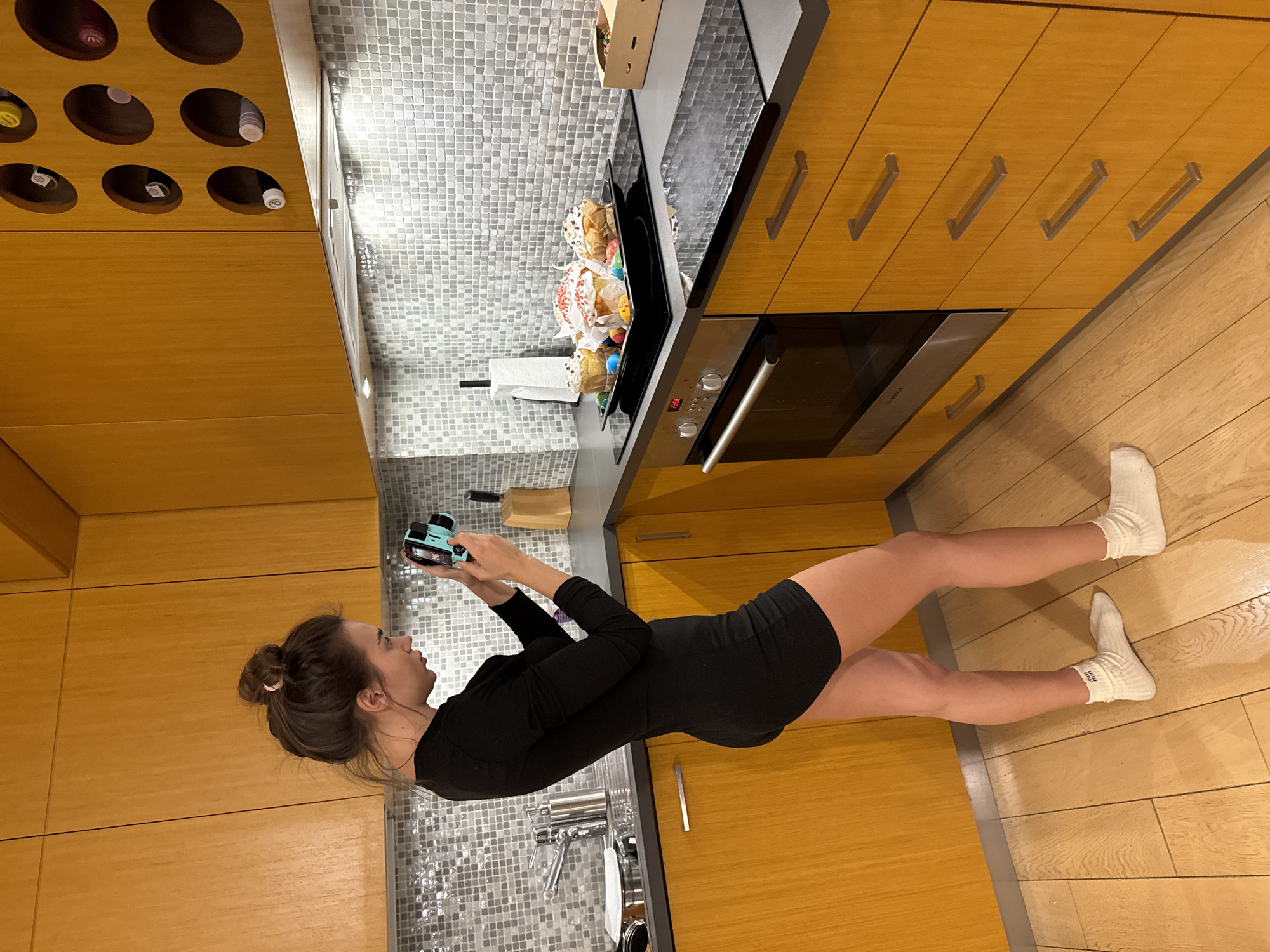 A woman in a black dress and white socks stands in a kitchen, holding a blue instant camera. The kitchen features light brown wood cabinets, a wine rack, a gray tiled backsplash, a black stovetop with Easter bread, and a built-in oven. The floor is made of light brown wood planks. The location is possibly a residential kitchen in Warsaw, Poland.