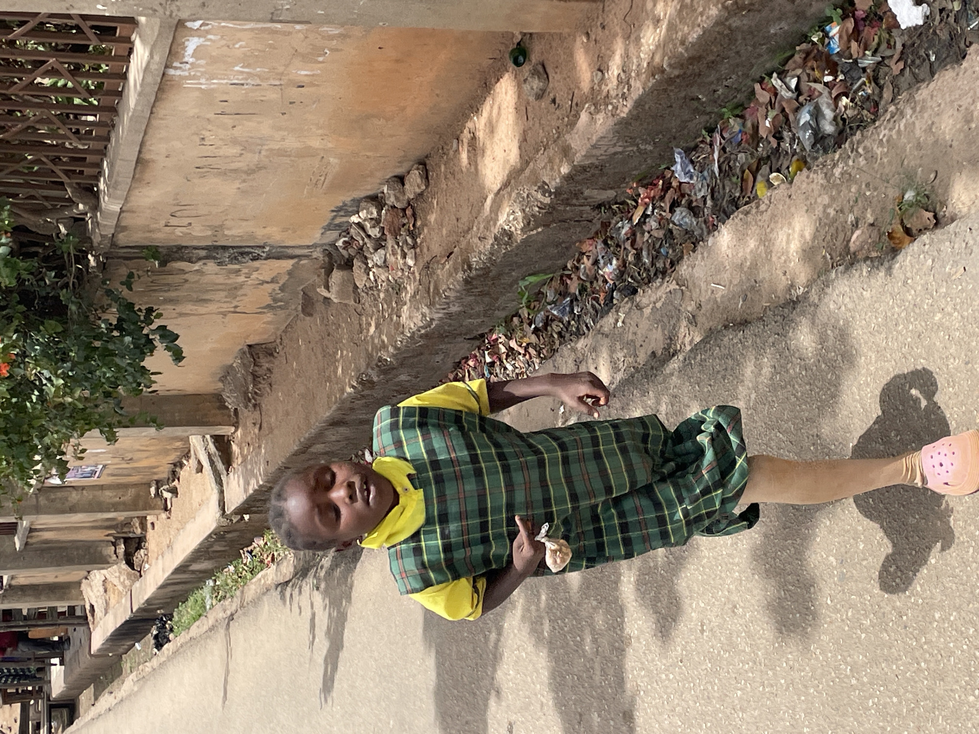 A young girl in a green plaid school uniform with yellow trim and pink shoes stands on a concrete sidewalk in Jos, Nigeria. Her shadow is cast on the pavement to her right. To her left, a concrete building abuts the sidewalk with a narrow ditch filled with litter separating it from the sidewalk. Trees and vegetation are present on the building side.