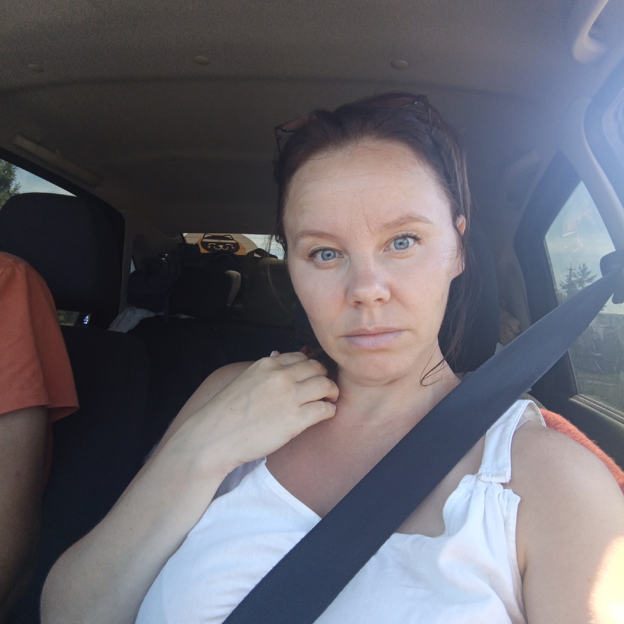 A medium shot captures a woman with light skin and blue eyes, wearing a white tank top, seated in the passenger seat of a car. Her dark brown hair is pulled back, and she is wearing sunglasses on her head. The woman is looking directly at the camera, with her right hand resting near her neck. A black seatbelt is diagonally across her chest. The interior of the car is visible, including the car's headliner and a window on the right side. In the background, through the window, a blurry view of trees and a cloudy sky is seen. The car appears to be in motion, indicated by the slight motion blur. The location is not in Cherkassy, Russia, as Cherkassy is in Ukraine.