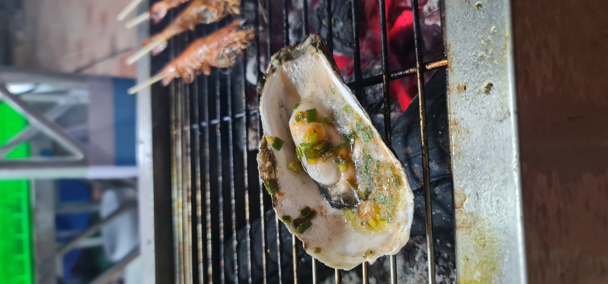 The image shows an oyster with green onions being grilled over hot coals on a metal grill. In the background, shrimp skewers are also grilling. The scene appears to be a street food stall in Đà Nẵng, Vietnam, given the commonality of grilling seafood in this city.