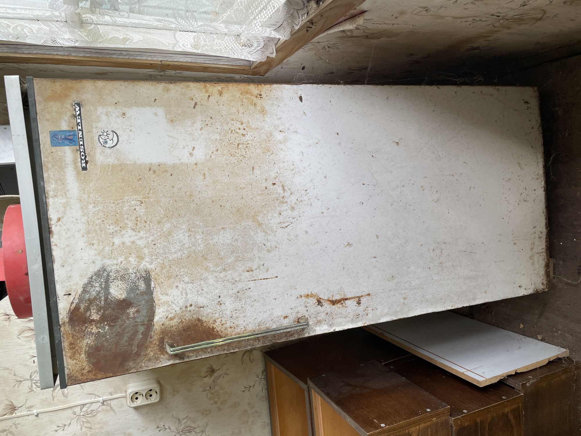 In an indoor, dimly lit scene of a residential room, a tall, white, and heavily rusted refrigerator stands against a floral-patterned wallpaper. The refrigerator door has a silver handle on the left side and two stickers towards the top right: one with a blue figure and the word "ROSENLEW" below it, and another smaller sticker featuring a crescent moon and stars. To the left of the refrigerator, a white electrical outlet is visible on the wallpaper. In front of the refrigerator and to its left are several wooden drawers or cabinet fronts, stacked and partially obscured. A white panel leans against the refrigerator's left side, near the bottom. The floor is made of concrete or dirt. To the right of the refrigerator, a wooden window frame with a white lace curtain is present. Cobwebs are visible in several areas, particularly in the corner between the refrigerator and the wall. The overall condition of the scene suggests neglect or disuse.