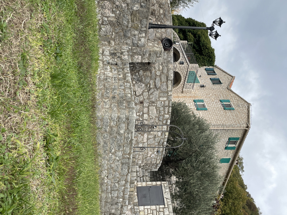 An outdoor scene in Đenjaši Česminovo, Montenegro, depicts a multi-story, light-colored stone building with a reddish-brown tiled roof situated on a hillside under an overcast sky. The building features multiple rectangular windows fitted with bright green wooden shutters; some are open, others are closed. A small balcony with a dark metal railing extends from the front of the house, supported by two stone arches.
In the midground, a substantial terraced stone wall, constructed from rough-hewn grey stones, occupies the lower and central portion of the image. A black, ornate streetlamp with two light fixtures stands on the upper left section of this wall. To its right, a dark, tire-like object rests on the stone surface. Further right, a black metal wellhead or decorative arch structure stands on another section of the stone wall, featuring a small bucket or pulley mechanism. To the far right, a series of stone steps are integrated into the wall, leading up to a dark grey metal door or panel set flush into the stone masonry.
A large olive tree with dense, silvery-green foliage is positioned between the stone wall and the building, partially obscuring the house. Denser green deciduous and evergreen trees cover the hillside behind and to the right of the house. The immediate foreground consists of unkempt grass, displaying varying shades of green and some drier, browner patches. No individuals, animals, or active interactions are present in the scene.