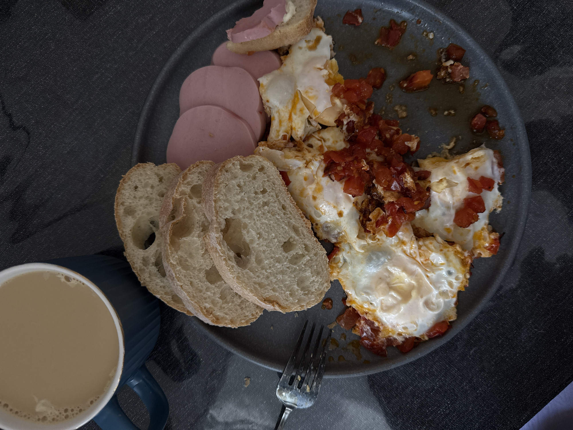 The scene depicts a meal, located in the city of Khimki, Russia, served on a dark grey, circular plate positioned centrally on a dark, textured fabric surface, possibly a tablecloth. The plate holds several components of a breakfast or brunch.
On the left side of the plate, three thick slices of light-colored, rustic bread, consistent with a ciabatta or similar artisan loaf, are stacked. To the upper right, multiple thin, circular slices of pale pink processed luncheon meat or sausage are arranged. One smaller piece of bread, partially consumed, is placed near the luncheon meat, topped with a spread of the same pink meat and a lighter, possibly mayonnaise or butter, substance.
The central and lower portions of the plate contain several fried eggs, some appearing intact while others are disturbed and mixed with finely diced red vegetables, likely tomatoes or red peppers, which exhibit signs of having been cooked. Residue from the eggs and vegetables, along with some liquid and crumbs, is visible on the plate, indicating the meal is partially consumed.
A blue ceramic mug with a ridged exterior and a subtle gold rim is visible in the upper left corner, containing a light brown beverage with small bubbles on its surface, consistent with coffee or tea mixed with milk. A silver-colored metal fork with four tines rests on the dark fabric to the left of the plate, extending slightly over its edge. The surrounding environment consists solely of the dark, patterned fabric surface. No individuals are visible.