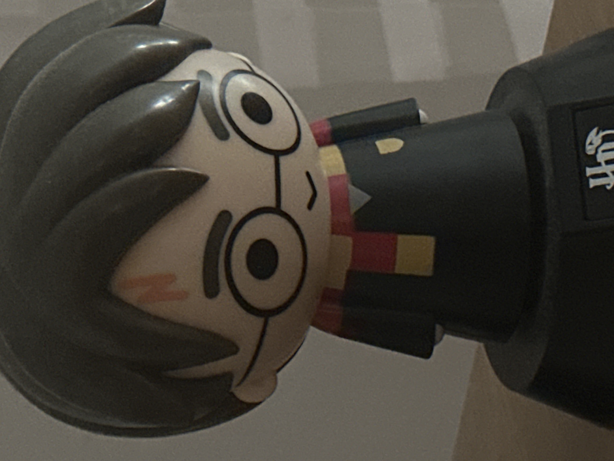 A close-up shot depicts a plastic toy figure of Harry Potter. The figure has dark gray hair, round glasses, a scar on its forehead, and a red and yellow striped scarf. The figure is attached to a dark gray base with white lettering on its side. The background is a light gray surface. There is no visible indication of the location in Budva, Montenegro.