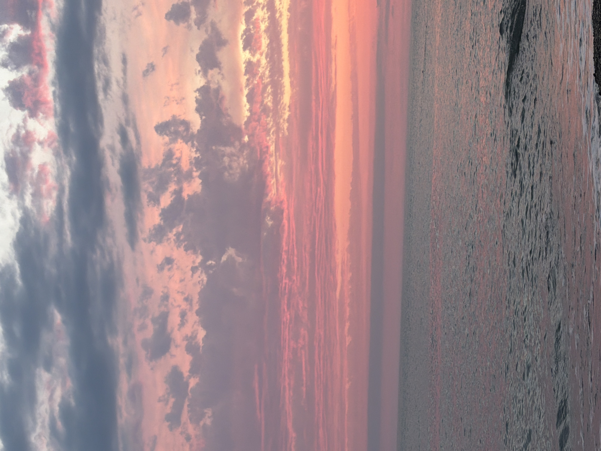 The image depicts a serene seascape at dusk. The sky is painted with a gradient of soft pinks and oranges from the setting sun, transitioning to shades of gray and purple in the upper clouds. Wispy clouds are scattered across the sky, some catching the light and appearing a vibrant pink. The ocean surface is calm, with gentle waves lapping at the shore, reflecting the colors of the sky. The horizon line is clearly defined between the sky and the sea. The lower portion of the image shows a narrow strip of dark, pebbled beach. There are no people or man-made structures visible in the scene. The overall atmosphere is peaceful and tranquil.