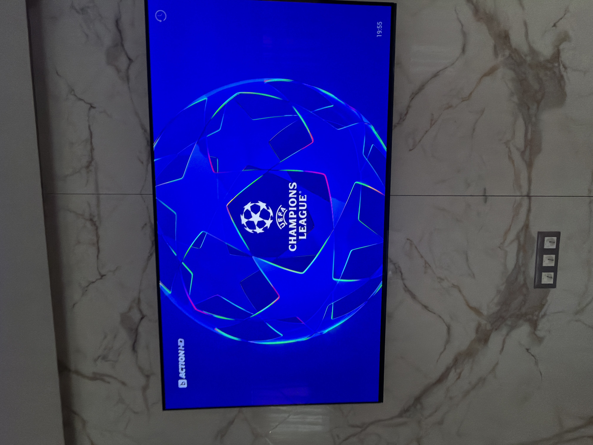 A television displaying the UEFA Champions League logo is mounted on a marble-patterned wall. The screen shows a blue background with the star-studded ball logo, with "UEFA CHAMPIONS LEAGUE" written vertically. "Action HD" is visible in the lower left corner, and the time "19:55" is in the upper right corner of the screen. To the right of the TV, on the wall, is a triple light switch. The location is unknown within the city of Asenovgrad, Bulgaria. No people are visible.
