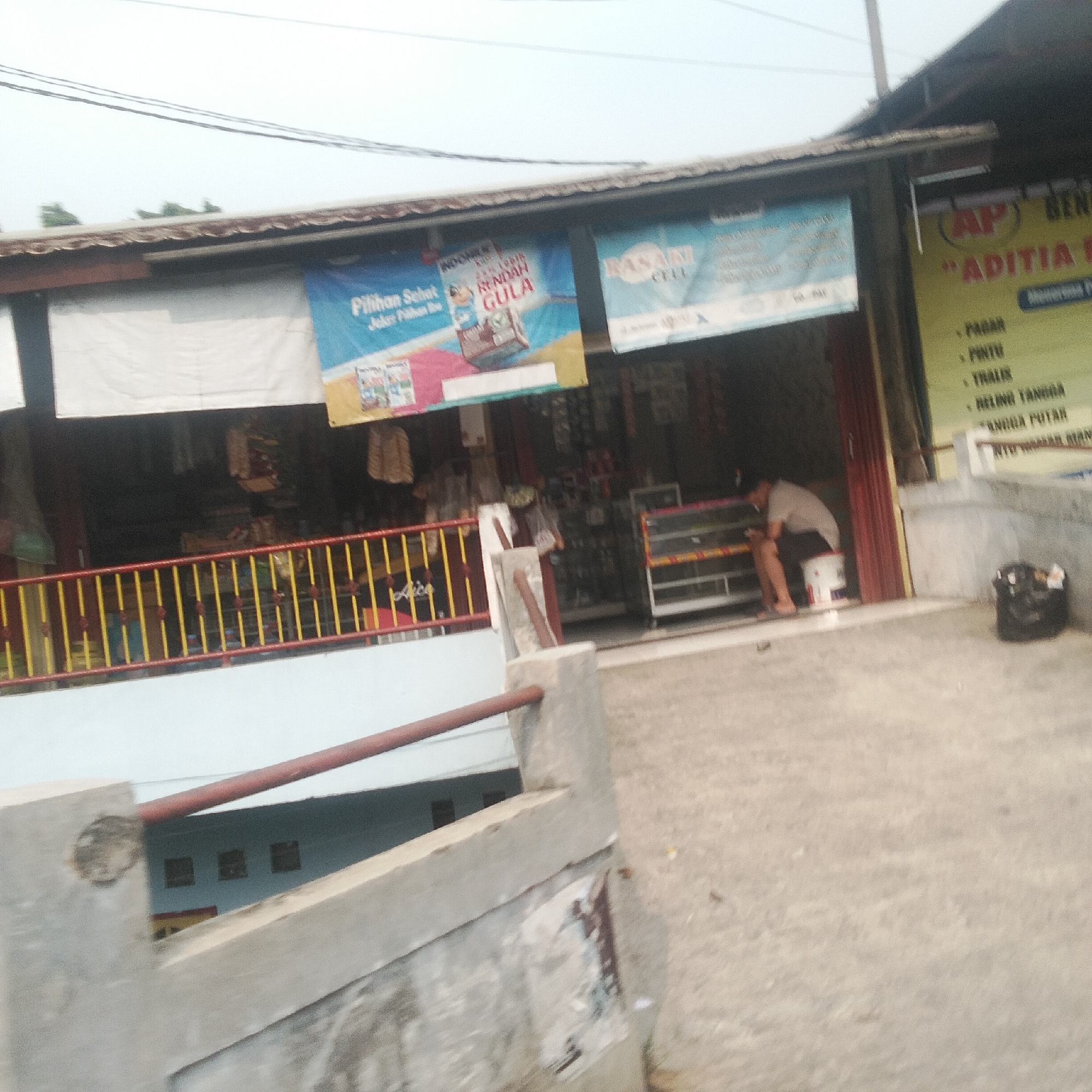 The image depicts the exterior of a shop in Bogor, Indonesia. The shop's facade features several banners, including one with the text "PILIHAN SEHAT" and "RENDAH GULA" in Indonesian, suggesting it offers healthy and low-sugar options. Another banner reads "RASALI CELL". A yellow and red railing runs along a walkway in front of the shop. Inside the shop, shelves are stocked with various items. A man is seated on a white bucket, facing away from the camera, near a display case. A black trash bin is visible on the ground outside the shop. The surrounding environment appears to be an urban setting, with buildings and power lines visible in the background. The overall image suggests a commercial street scene in Bogor.