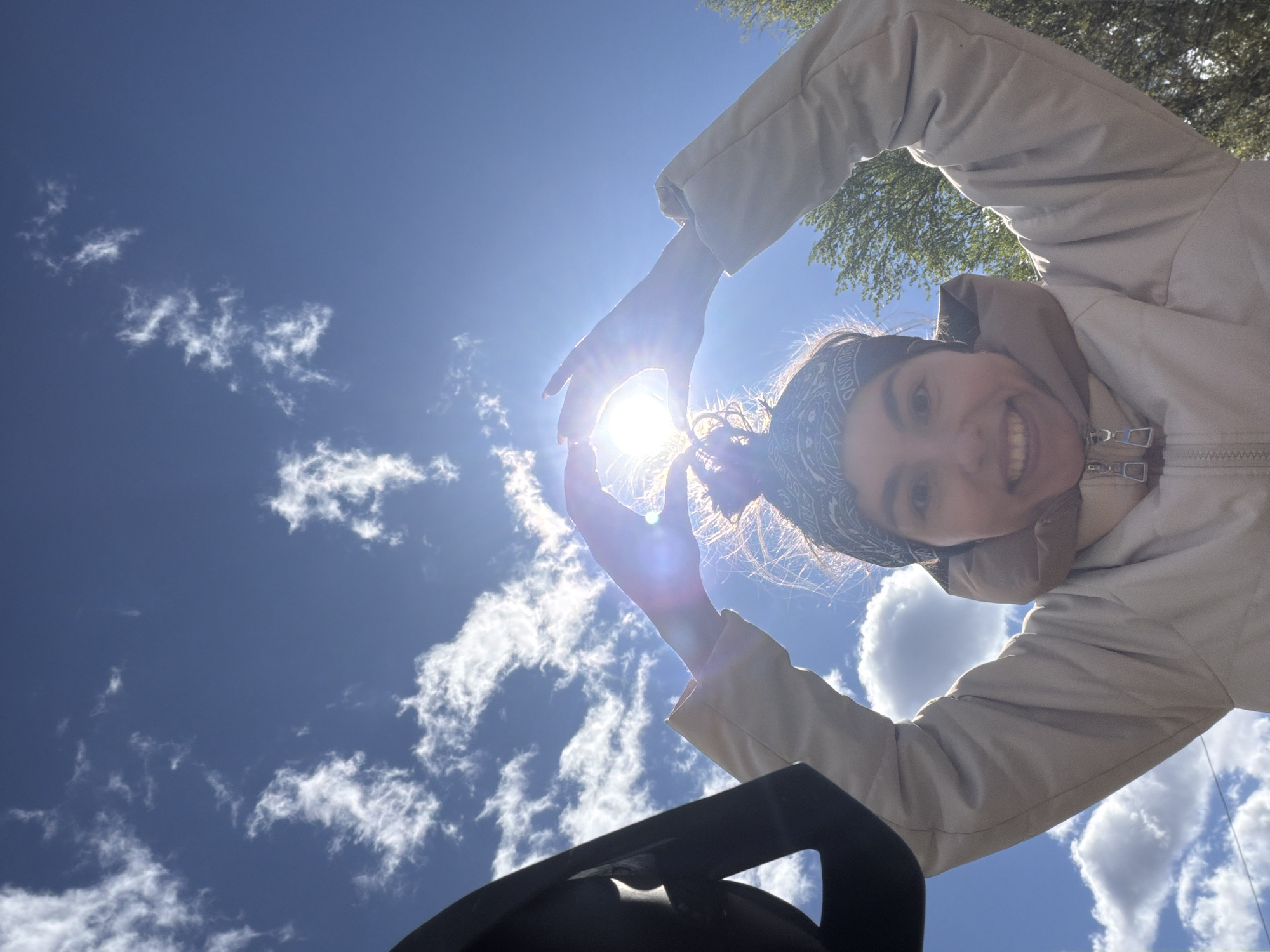 A low-angle shot captures a person against a blue sky with scattered clouds. The person, who is smiling, is wearing a light-colored jacket and a patterned headscarf. Their arms are raised, forming a heart shape around the sun. A black, undefined object is visible in the lower portion of the frame. The location of the image is unknown.