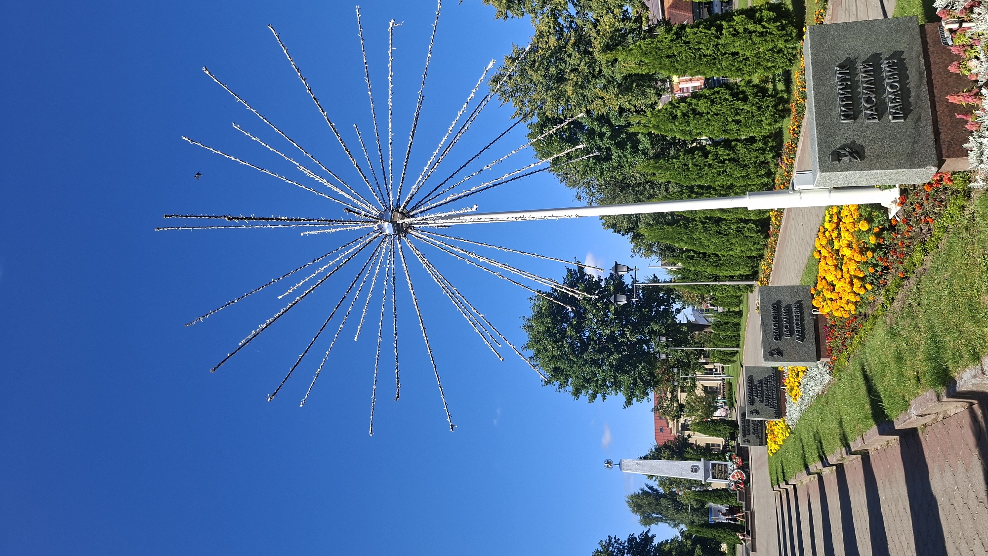 The image depicts a memorial site in the city of Pastavy, Belarus, under a clear blue sky. In the foreground, a large, starburst-shaped light fixture with numerous thin, metallic rods extends outwards from a central pole. In front of this structure are several granite plaques inscribed with names, set amidst vibrant flower beds of yellow and orange marigolds, as well as some red and pink flowers. A narrow pathway with red brick edging runs parallel to the flower beds. Further back, a tall, obelisk-shaped monument stands prominently, also with inscriptions and possibly wreaths at its base. A paved walkway, lined with green trees and manicured cypress trees, leads towards the monument. Streetlights are visible along the pathway. In the far background, buildings with red roofs and a few distant trees are discernible. One person is partially visible walking on the paved walkway in the middle ground to the left of the obelisk.