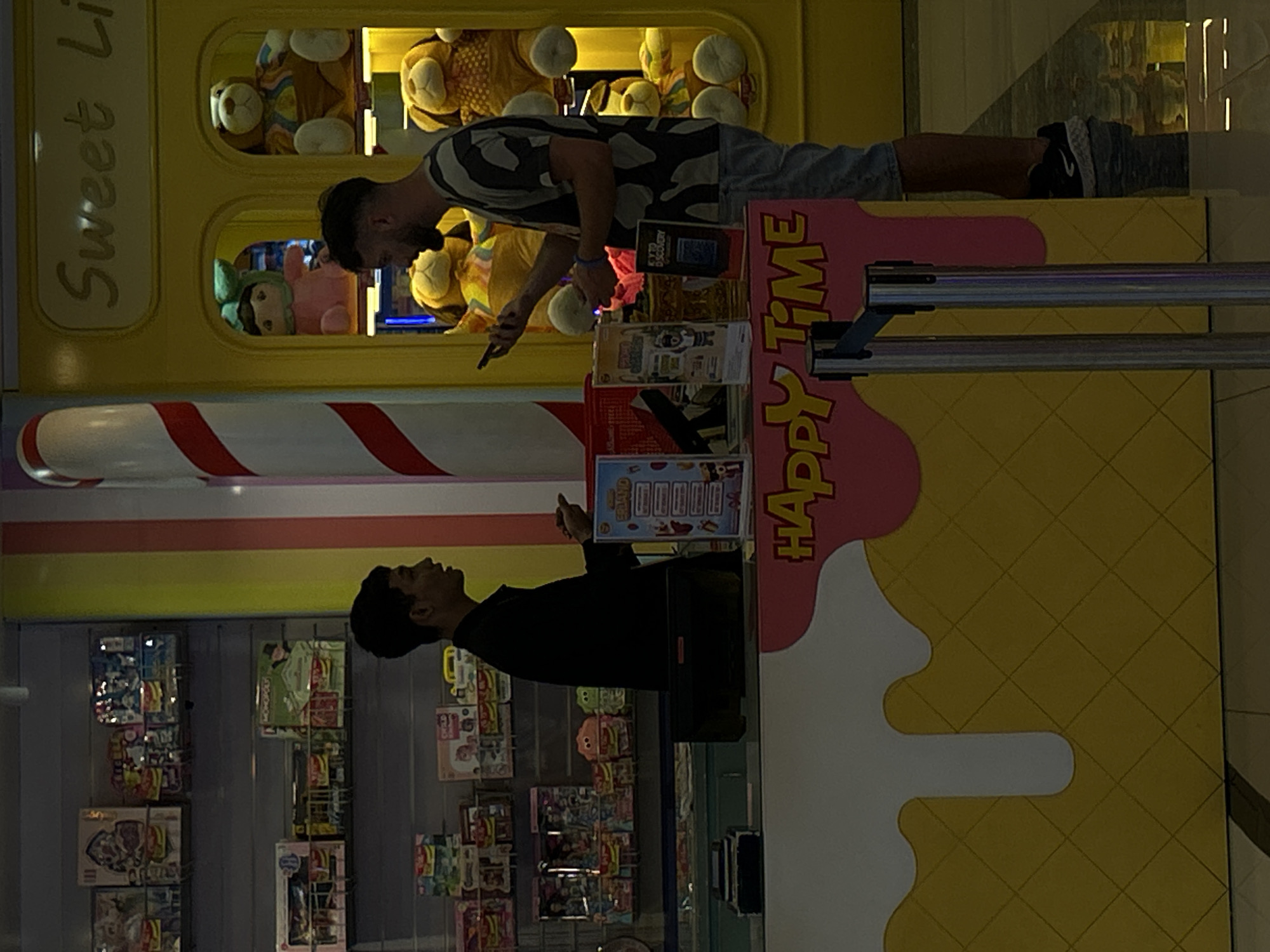 In Kuta, Indonesia, an employee wearing a black shirt stands behind a counter in a brightly lit store. The counter is decorated with a pink and yellow "Happy Time" graphic and has several merchandise displays on it. A customer, wearing a camouflage t-shirt and denim shorts, stands in front of the counter, looking at a mobile phone. Behind the counter, shelves are stocked with various toys and merchandise. To the right of the counter, a display cabinet filled with large stuffed animals is visible, with a "Sweet Life" sign above it. The store appears to be a toy or candy shop, possibly located within a mall or entertainment complex.