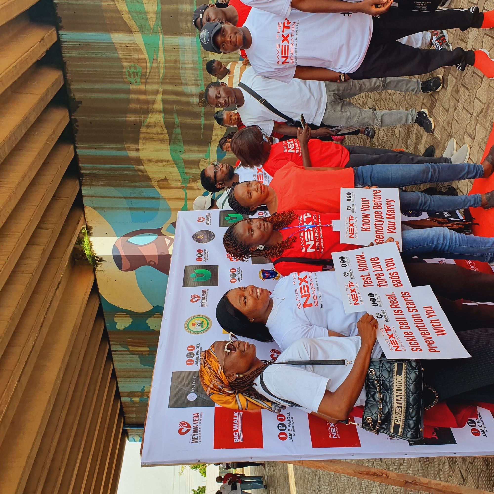 This image depicts an outdoor event, possibly a health awareness campaign, taking place in Jos, Nigeria, as indicated by the presence of the "GOVERNMENT OF PLATEAU STATE OF NIGERIA" logo on a banner. A group of approximately 15 to 20 people, diverse in age and gender, are gathered. The foreground is dominated by a large white banner with red and black lettering and various logos, including "THE MEYIWA VERA EMPOWERMENT FOUNDATION," "ScoutsCity," and "JAMIE PAJOEL INTERNATIONAL." Several individuals are holding white signs with red text. One sign reads, "SAVE THE NEXT GENERATION. Our parents didn't know but now we know." Another sign states, "Sickle Cell Is Real. Prevention Starts With You." A third sign says, "Test. Know. Before Love leads You." Another sign asks, "Know Your Genotype Before You Marry."
The people are dressed casually, with many wearing t-shirts, some red and some white, that also feature the "SAVE THE NEXT GENERATION" slogan. Some individuals are smiling and looking towards the camera, while others appear to be engaged in conversation or observing the surroundings. One woman in the foreground is wearing a yellow headscarf and sunglasses and carrying a black Christian Dior handbag.
The background reveals a concrete structure, likely an underpass or bridge, with a painted mural on the wall. The mural features abstract shapes and colors in shades of blue, yellow, brown, and green, with elements resembling clouds and possibly a stylized human figure. The ground is paved with interlocking bricks. The overall atmosphere appears to be that of a community gathering or event.