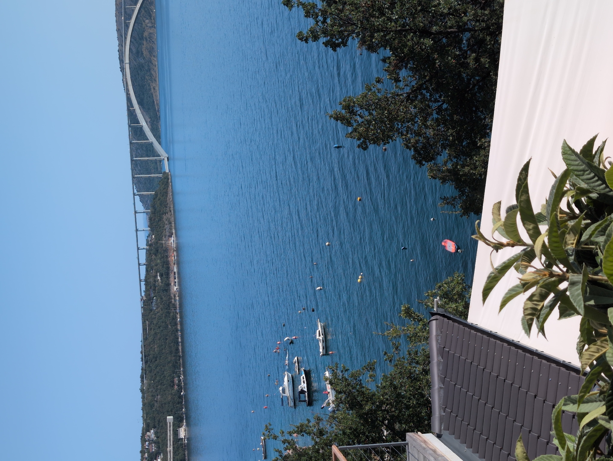 The image depicts a serene coastal scene in Kraljevica, Croatia, with a prominent bridge spanning across the blue water in the background. Several boats, including a jet ski, are anchored in the bay, with buoys marking their positions. Lush green trees frame the foreground, and part of a tiled roof and white awning are visible, suggesting a residential or hospitality setting. The sky is clear and blue, indicating a sunny day. The Krk Bridge, a significant landmark connecting the island of Krk to the mainland, dominates the horizon. The water appears calm, reflecting the clear sky.