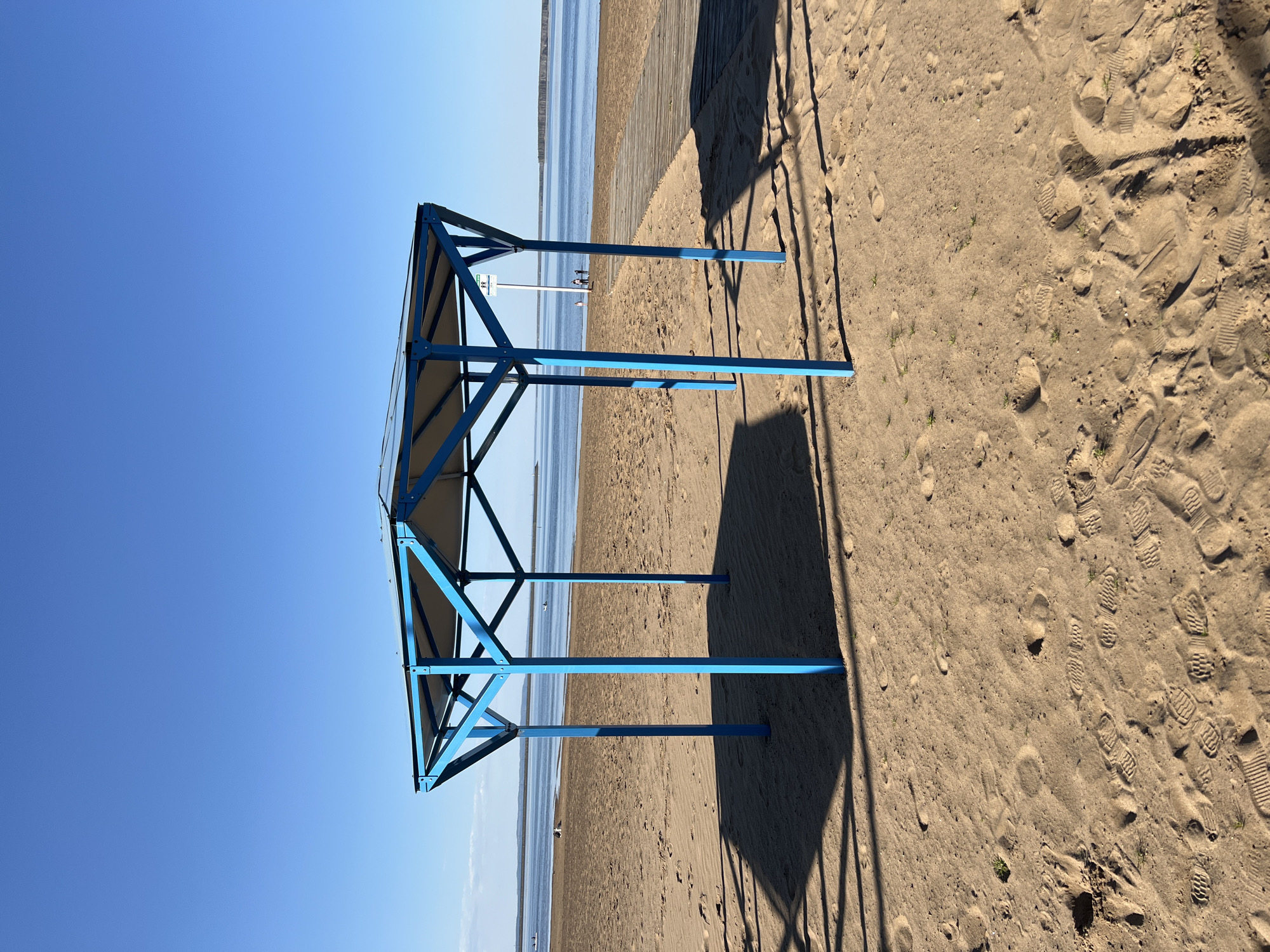 The image shows a blue metal structure, possibly a shade or shelter, on a sandy beach next to a body of water under a clear blue sky. Footprints are visible in the sand. The location is likely Chaikovsky, Russia. There are people visible in the distance. A boardwalk is on the right side of the image.