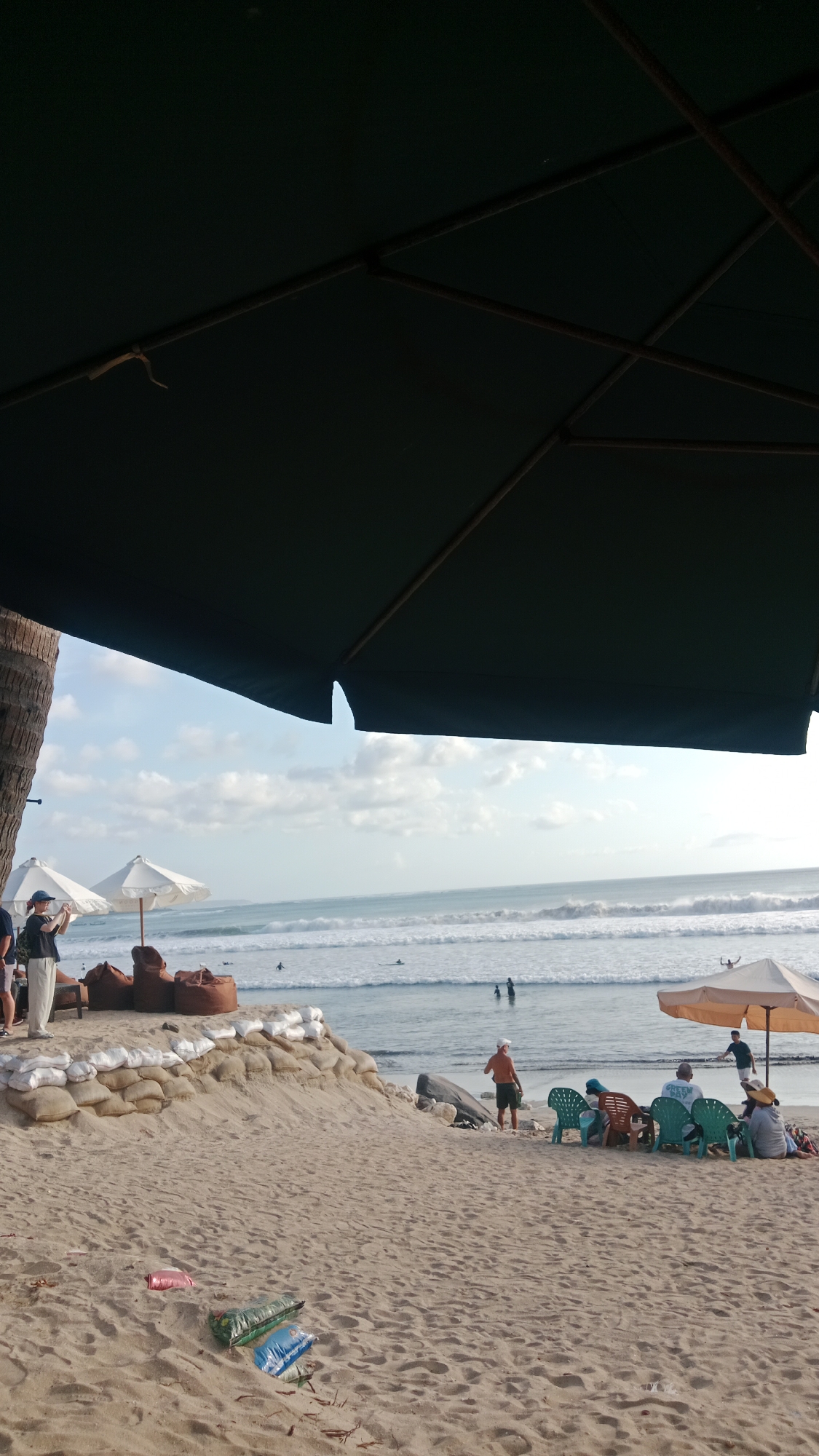 The image depicts a scene on the beach in Kuta, Indonesia, under a large dark green umbrella. In the foreground, the sandy beach is strewn with some litter, including colored plastic bags and debris. A small sand barrier, reinforced with sandbags, is visible. Behind the barrier, several people are seated in colorful chairs under a beige umbrella, facing the ocean. Some of these individuals appear to be tourists. Further back, two white beach umbrellas are set up, with large beanbag chairs nearby. A person is standing near one of the white umbrellas, holding a camera or phone. In the ocean, waves are breaking, and a few surfers can be seen in the water. The sky is partly cloudy.