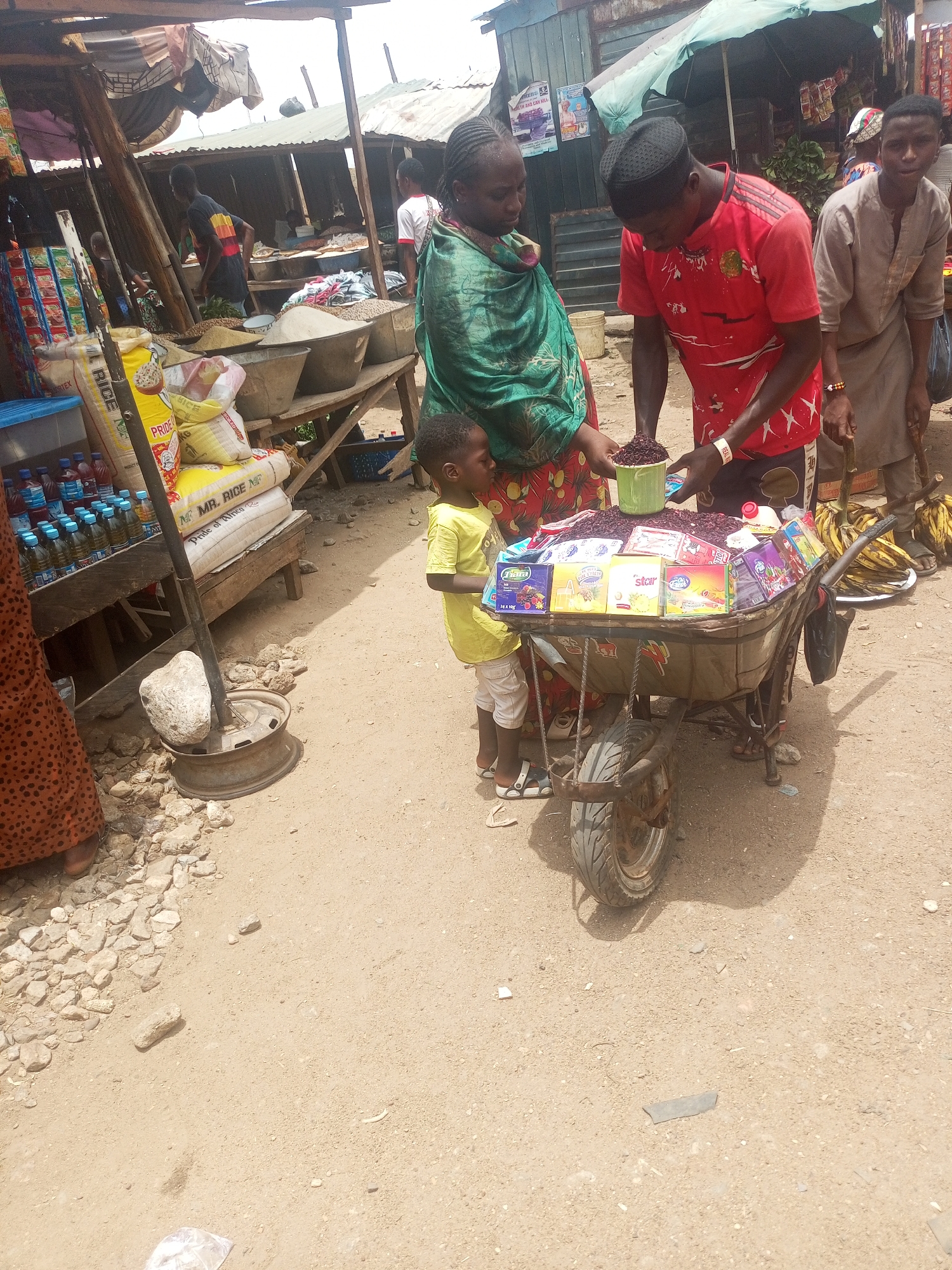 The image captures a scene at an outdoor market in Sabon Tasha, Nigeria. In the foreground, a man in a red shirt is selling dried hibiscus flowers (zobo) from a wheelbarrow. The wheelbarrow also contains small boxes of what appear to be tea or beverage mixes. A woman in a green wrap stands beside the wheelbarrow, seemingly interacting with the vendor. A young child in a yellow shirt is close to the wheelbarrow, possibly accompanying the woman. Other people are visible in the background, suggesting a bustling market environment with various stalls selling goods such as rice and other provisions. The ground appears to be a mix of sand and gravel.