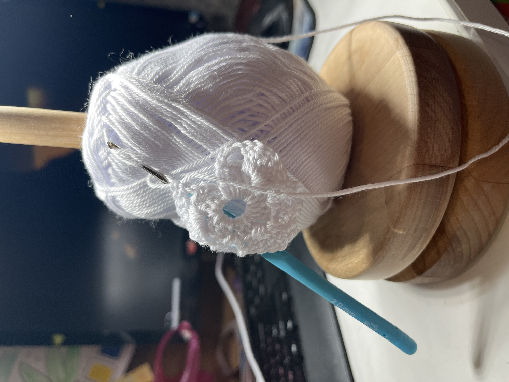 The image shows a close-up of crocheting supplies. A ball of white yarn sits on a wooden yarn holder, with a partially crocheted white circle emerging from the ball. A blue 2.0 mm crochet hook rests near the crocheted piece. A brown wooden dowel is inserted through the yarn ball's center. Two silver stitch markers are attached to the yarn ball. The background shows a computer screen and other blurred objects on a table. The location cannot be determined from the image alone.