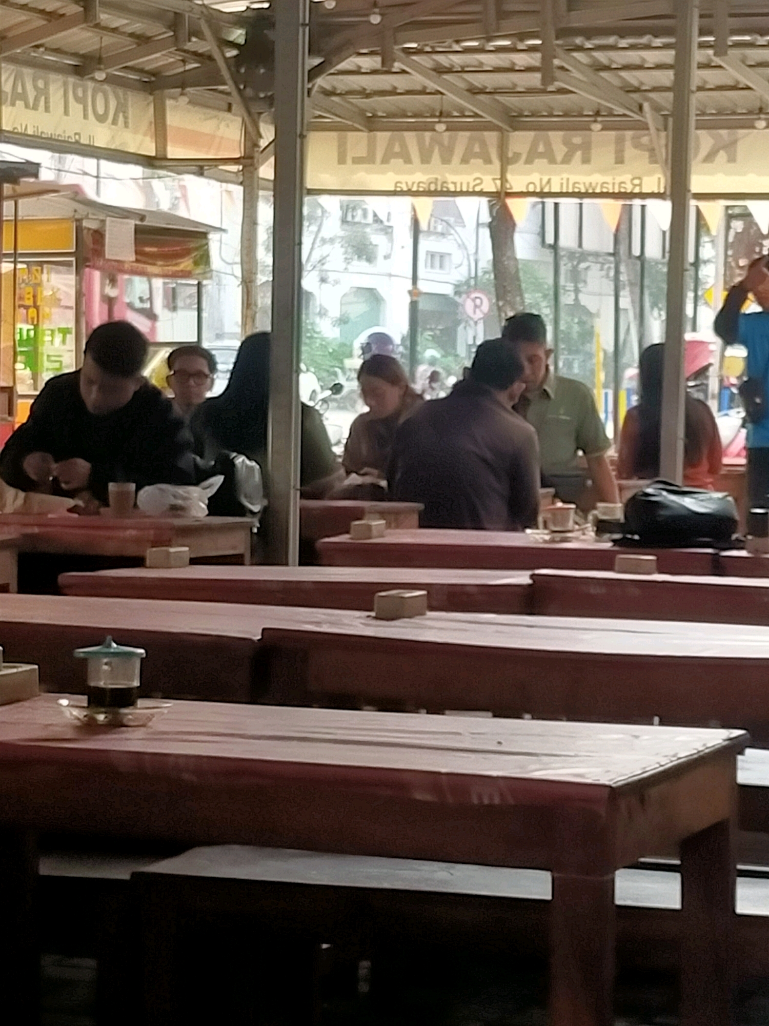 This is an image taken from inside a covered outdoor seating area of a cafe or restaurant, likely in Surabaya, Indonesia, judging by the text on the signage. The signage above reads "KOPI RAJAWALI" which translates to "Eagle Coffee". The interior is open-air, with a metal roof supported by beams. Several wooden tables and benches are arranged in the seating area. In the foreground, a small glass cup with a dark liquid, possibly coffee or tea, sits on a saucer on a table.
Several people are visible in the mid-ground and background. On the left, a man in a dark shirt is seated at a table, appearing to eat or prepare food. To his right, a woman wearing glasses is seated. Further back, other individuals are seated at tables, some with drinks and food. A person wearing a bright blue shirt is visible on the far right, partially cut off by the frame. The background shows a street scene with buildings and trees, suggesting an urban environment. The overall impression is of a casual dining establishment.