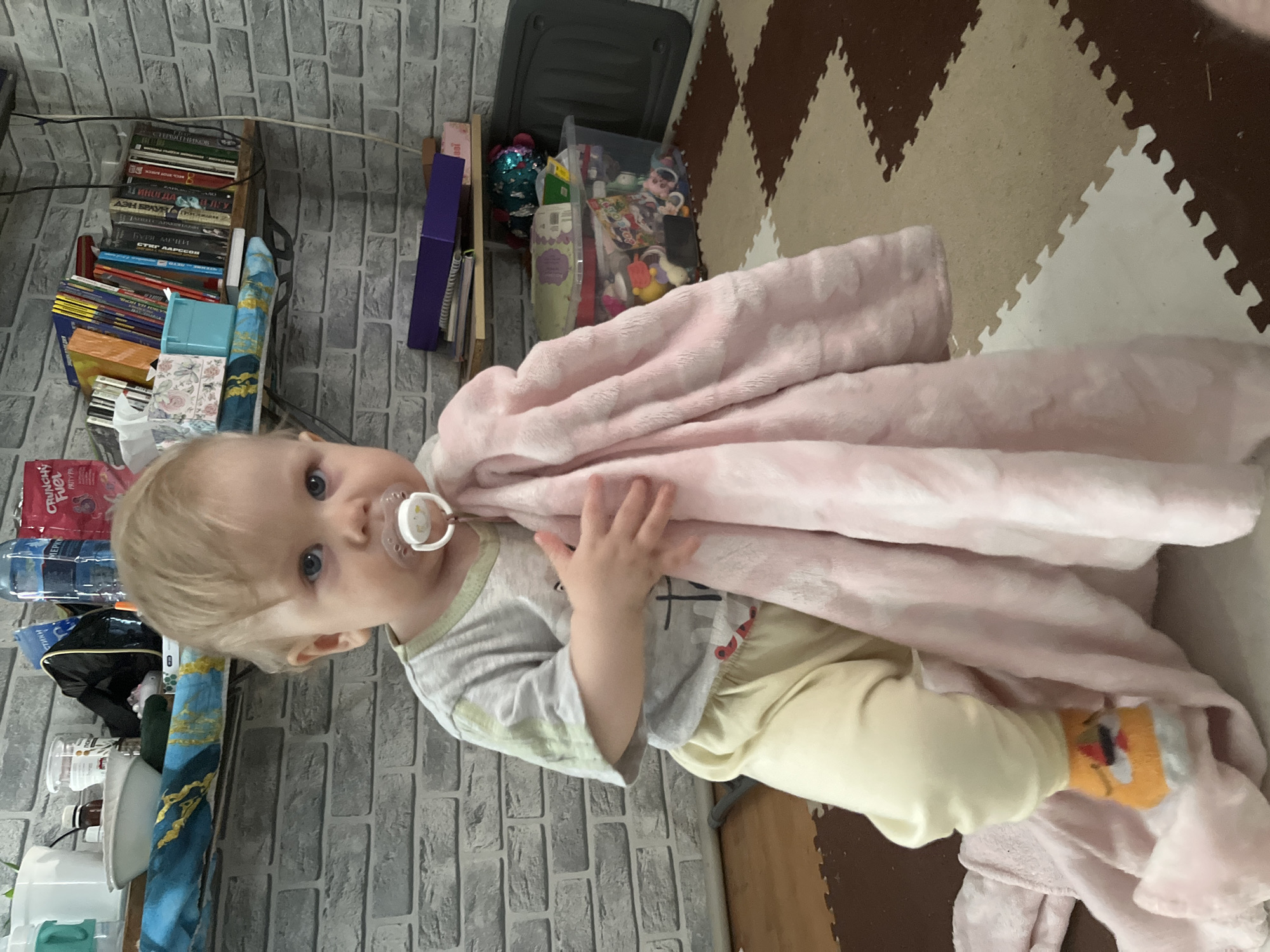 An indoor, eye-level shot captures a young child, likely a toddler, with fair skin, light blonde hair, and blue eyes, looking directly at the camera. The child wears a light-colored short-sleeved top, cream-yellow pants, and a white pacifier in their mouth. They hold a soft, light pink blanket, which drapes over their right arm and extends downwards, partially obscuring the lower body. One foot, partially visible, wears an orange sock with a cartoon character design.
The background features a wall adorned with grey brick-patterned wallpaper. To the left of the child, a wooden shelf holds numerous books, some with visible Russian titles such as "ГЕРОИ ТЕНИКОВ" (Heroes of Shadows), "ОТЦЫ И ДЕТИ" (Fathers and Sons), and "БУРЯ МЕЧЕЙ" (Storm of Swords), alongside "СТИВ ЛАРССОН" (Stieg Larsson). Various household items, including bottles (one labeled "CRUNCHY FUEL"), are also on the shelves. To the right, part of a soft play mat with interlocking brown, beige, and white zig-zag patterns is visible on the floor. Behind the blanket, a clear plastic storage bin filled with toys and a dark grey storage box are present. The child appears calm and observant. The Russian text on the books suggests the scene is set in Vesyoloye, Russia.