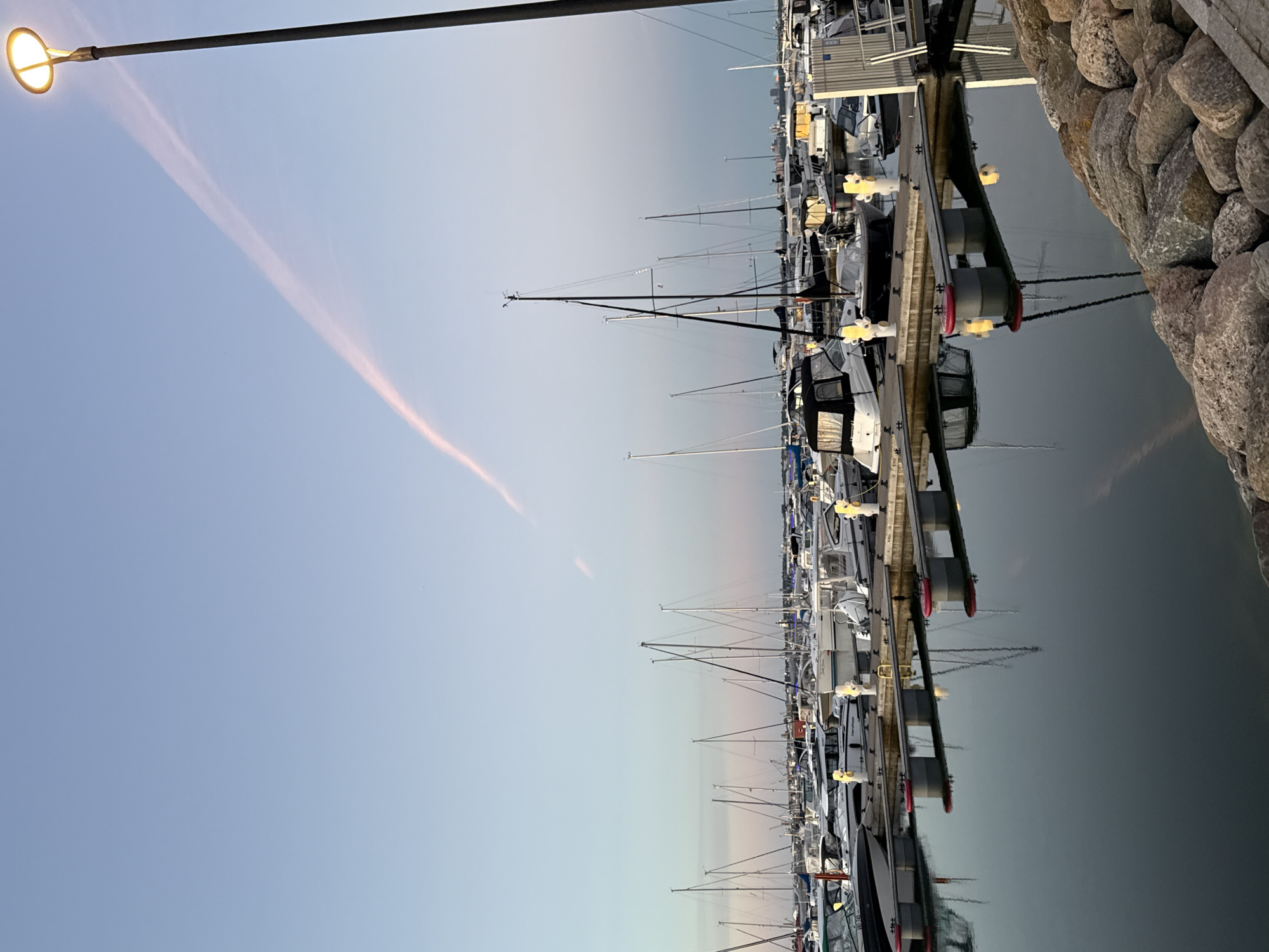 The image depicts a marina at dusk, likely located in Tallinn, Estonia. The sky is a soft gradient of blue and pink, with a wispy pink cloud formation stretching across it, illuminated by a street lamp on the right side of the frame. Rows of moored sailboats and motorboats are visible, their masts and hulls casting reflections on the calm water. The boats vary in size and design, some covered with tarps. The marina infrastructure, including docks and bollards, is also present. The foreground features a rocky embankment. No people are visible in the scene.
