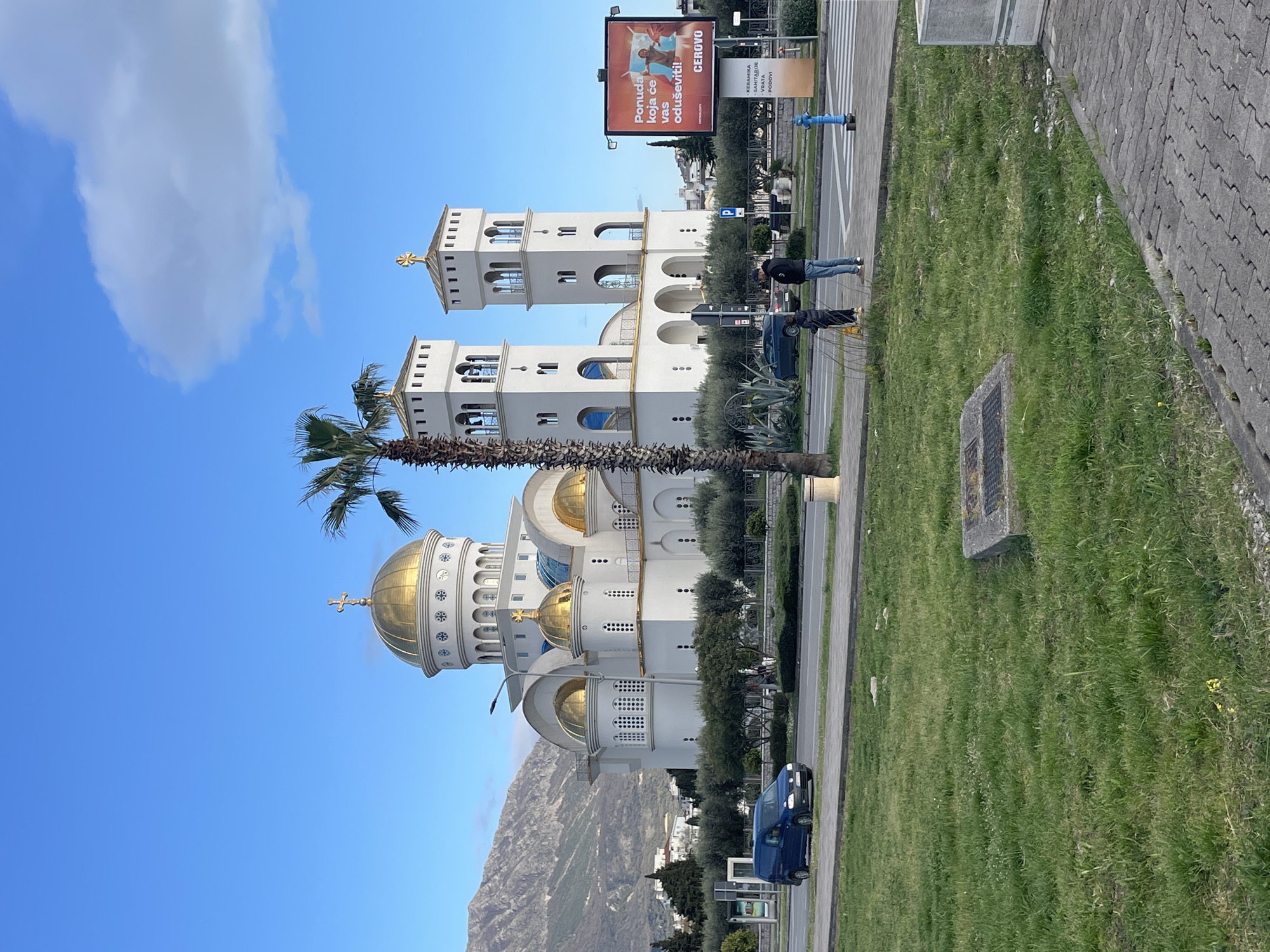 The image shows the Church of St. Jovan Vladimir in Bar, Montenegro. The church is white with golden domes and two bell towers. A tall palm tree stands next to the church. In the background, a mountain range is visible. In the foreground, there's a green grassy area bordering a sidewalk. A road runs parallel to the church, with a dark blue vehicle parked along it. Two people are standing on the sidewalk near a billboard advertising CERVOVO. A blue fire hydrant is also visible on the sidewalk.