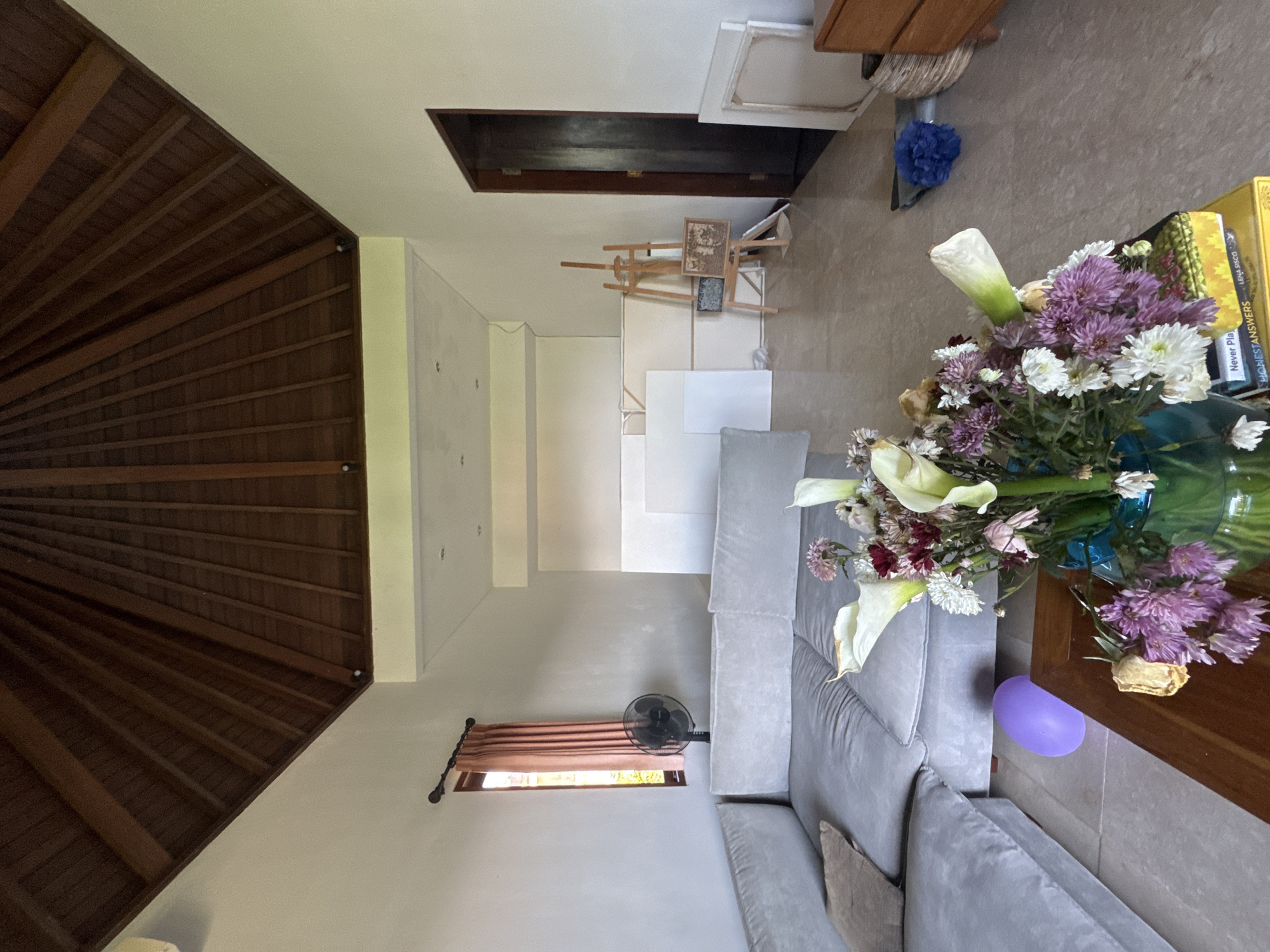 The image shows the interior of a room in Kuta Selatan, Indonesia. The room has a wooden ceiling and light-colored walls. There is a staircase leading upwards. A gray sofa is positioned in the lower center of the frame. A flower arrangement in a vase sits on a wooden table next to the sofa, and a purple balloon is visible. The room has a window with a pink curtain and a fan. Art supplies, including an easel and canvases, are present in the background.