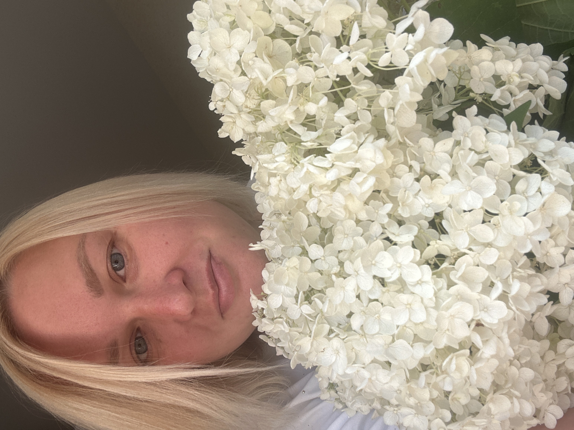 A blonde woman is holding a large bouquet of white hydrangeas. She is looking towards the camera, and her face is partially obscured by the flowers. The background is dark, suggesting an indoor setting. The image is a close-up shot, with the woman and the flowers filling most of the frame. The exact location in Chaikovsky, Russia, is not discernible from the image.