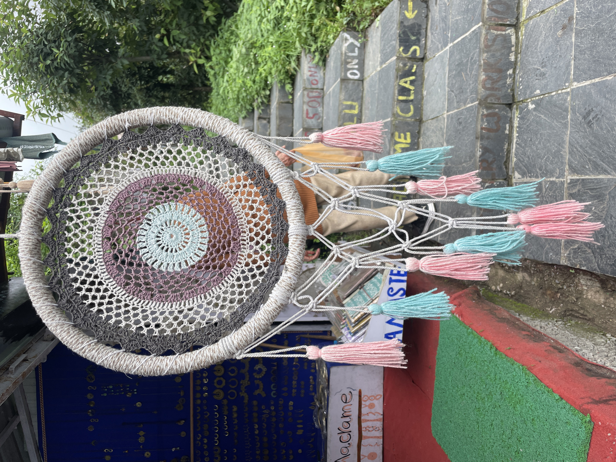 A close-up, low-angle, full shot captures a handmade dreamcatcher with white, gray, purple, and light blue crocheted concentric circles, hanging in front of a stall in Bhagsu, India. The dreamcatcher features white rope wrapping around its outer hoop and is adorned with multiple pink and turquoise tassels. To the left, a red platform displays a green artificial grass mat and a sign with the word "Macrame" written on it. Behind the dreamcatcher, a person wearing an orange sweater and tan pants is partially visible, holding a yellow bag. In the background, a blue display wall with various trinkets and jewelry hangs, along with stone stairs that ascend upwards. Yellow graffiti is visible on the stairs, with some words readable as "500 ONLY" and "ME CLASS". Lush green trees and foliage frame the scene, suggesting an outdoor environment in a hilly or mountainous region. The overall impression is one of a vibrant, artisan marketplace.