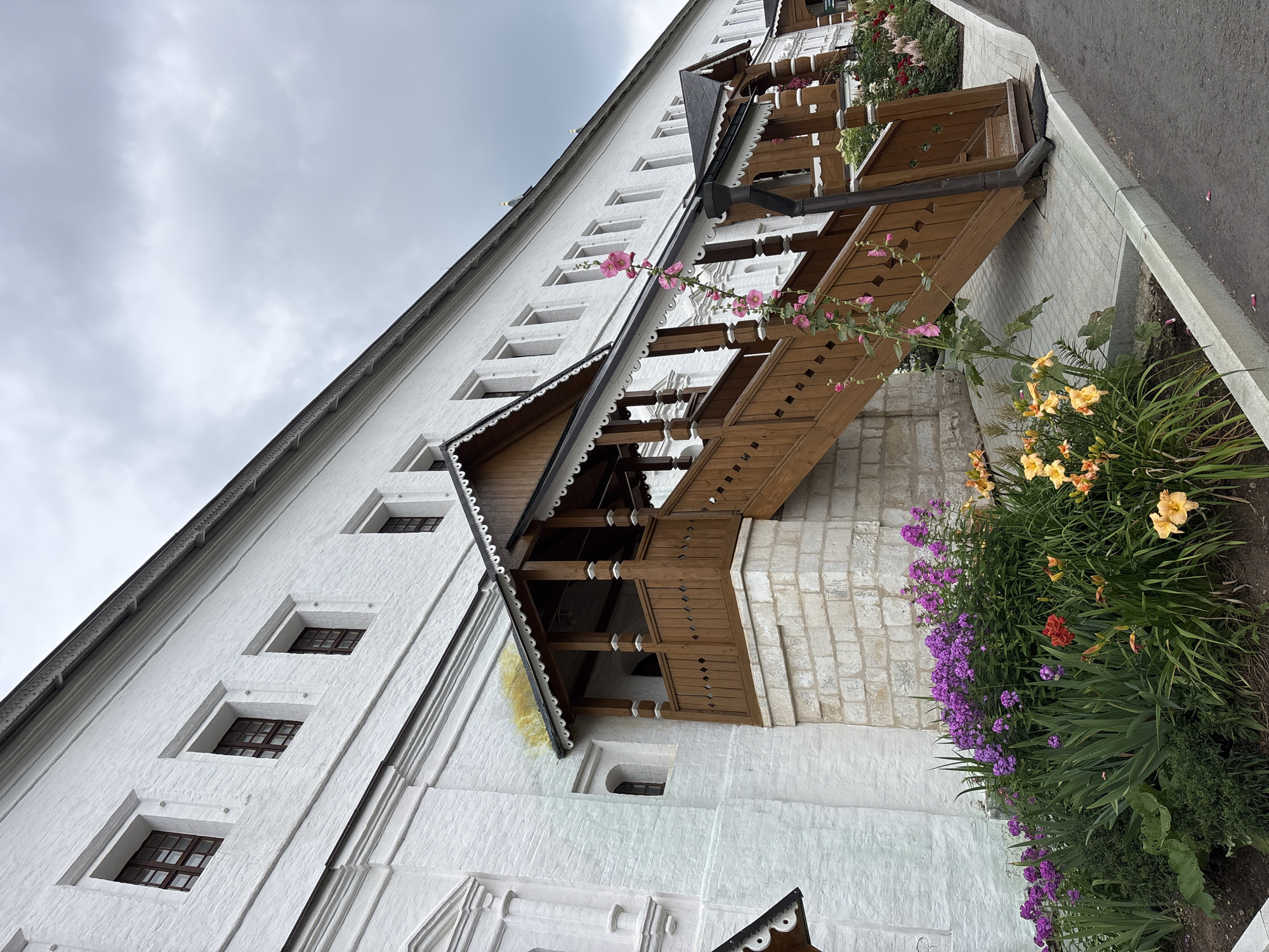 The image depicts a view of the Savvino-Storozhevsky Monastery in Zvenigorod, Russia. A white brick building with numerous windows stretches horizontally across the frame. In the foreground, a wooden staircase with intricate carvings leads up to an entrance, flanked by a flowerbed bursting with purple, pink, and orange blooms. Pink hollyhock flowers climb the side of the staircase. The sky is overcast and gray. The ground is paved with gray interlocking stones.