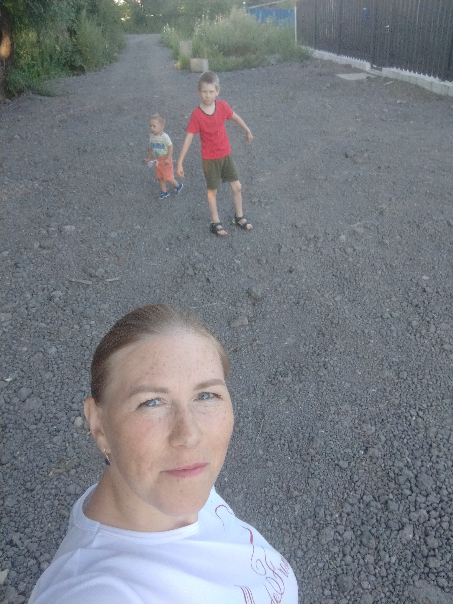 A selfie-style photo shows a woman in the foreground with freckles on her face, wearing a white t-shirt. Behind her, two boys are on a gravel path. The older boy, wearing a red t-shirt and dark shorts, stands slightly to the right. To his left and further back, a younger boy in an orange and striped shirt runs. The path is flanked by greenery and a dark fence on the right. The scene appears to be outdoors in Komsomolsk-on-Amur, Russia, possibly a residential area or park.