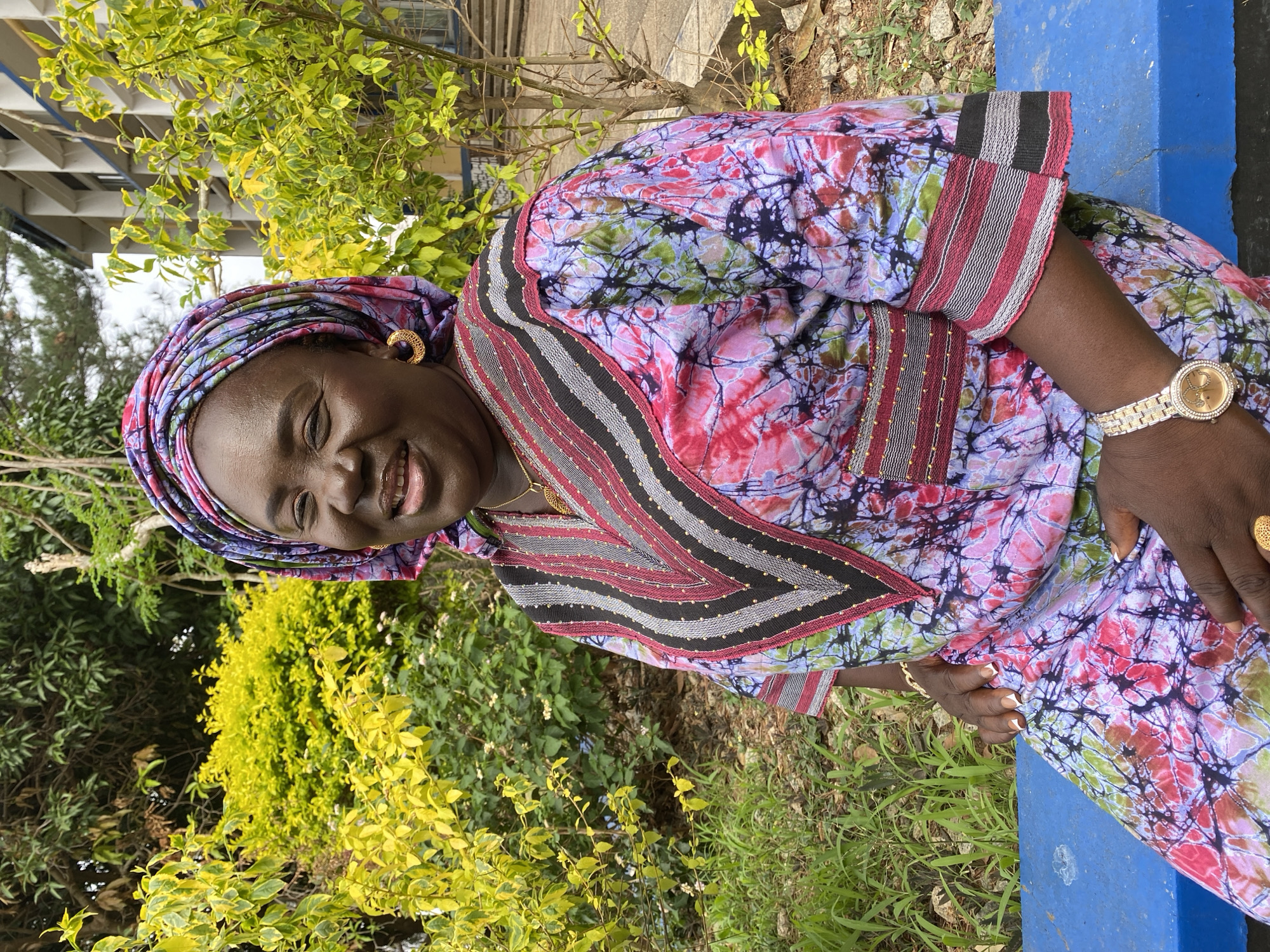 A smiling woman is captured in an outdoor setting in Bukuru, Nigeria. She has dark skin and is looking directly at the camera with a broad, joyful smile that reveals her teeth. Her head is adorned with a colorful headwrap, matching her vibrant, patterned garment, which appears to be a dress or top. The fabric features a batik or tie-dye style with a mix of pink, purple, green, and black. The garment has distinctive striped cuffs and a V-neckline detailed with red, black, and grey stripes, accented by small gold beads.
She is wearing gold-colored hoop earrings, a delicate gold necklace, and a gold-toned watch with a sparkling bezel on her left wrist. Her fingernails are visible, featuring white French tips. The woman appears to be seated on a blue bench or structure, part of which is visible to her right. The background is filled with rich green and yellow-green foliage, suggesting a lush garden or park environment. Natural daylight illuminates the scene, indicating it is daytime. No discernible text is visible in the image.