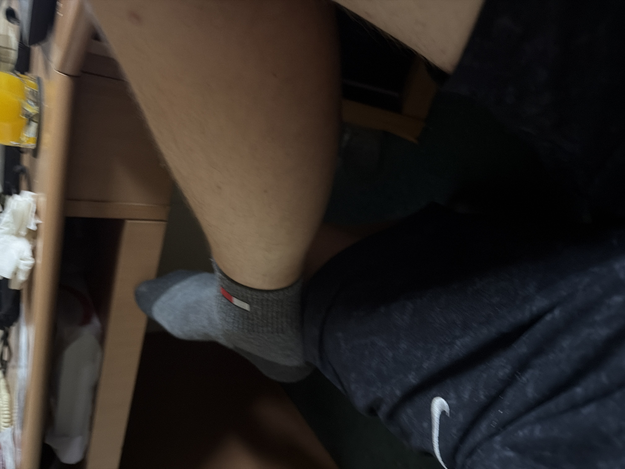 The image shows a close-up, low-angle view of a person's legs. The person is wearing a grey sock with a red and white stripe detail and dark patterned shorts. The legs are resting on a dark surface, possibly a chair or the floor. In the background, a wooden desk is visible, with some objects on it, including a glass of yellow liquid and some white items that appear to be tissues. The scene is indoors, likely in a domestic or office setting. There is no information in the image to suggest the location is Tuzla, Turkey.