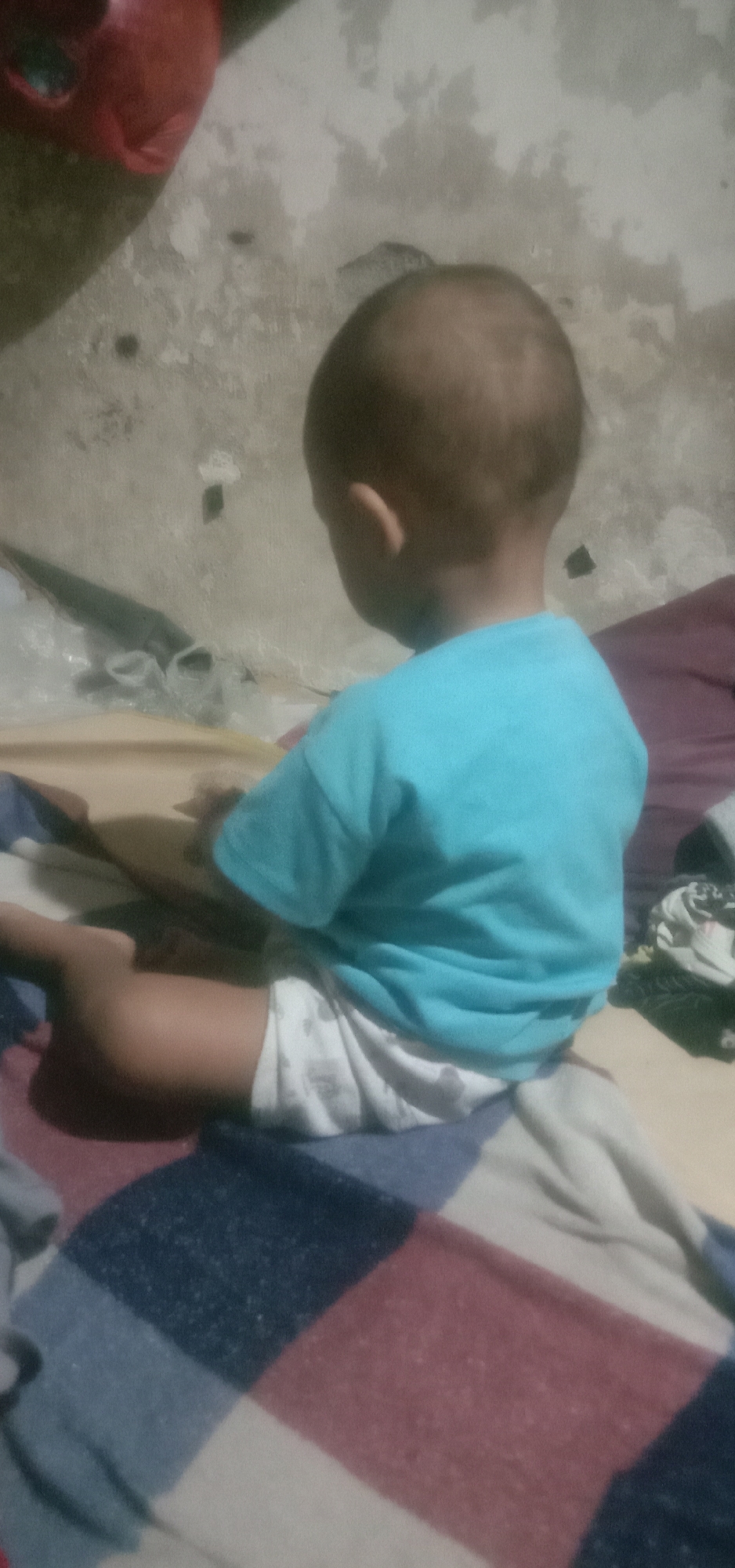 The image shows a young child from behind, sitting on a multi-colored blanket. The child is wearing a light blue t-shirt and a white diaper. The background appears to be a textured wall. To the left of the child, a red object is partially visible. The scene is indoors and appears to be a residential setting, possibly in Banjar Dowo, Indonesia.