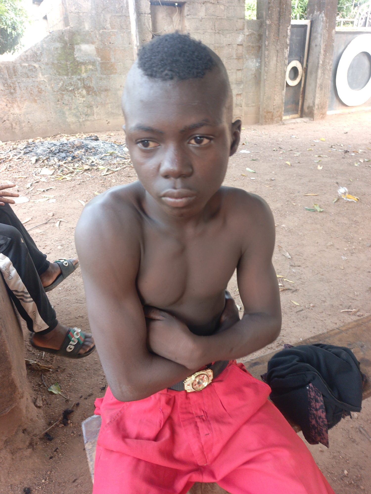 A shirtless young person is sitting on a wooden bench, wearing red pants with a gold-colored belt buckle. The person has their arms crossed. Next to the person is a black jacket. Another person is partially visible on the left, wearing pants with a white stripe and sandals. The ground is dirt. In the background is a wall, a pile of ash, and a gate with circular white decorations. The setting is in Akwanga, Nigeria.