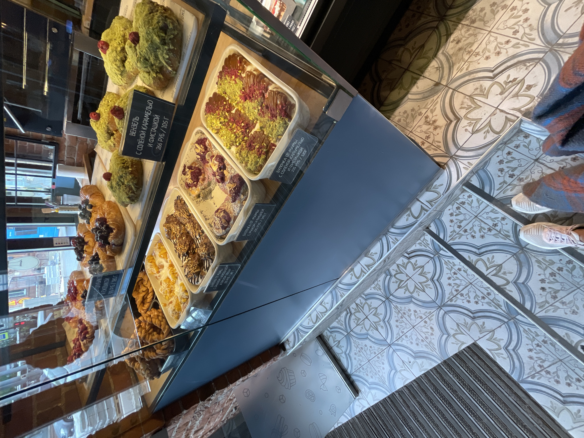 The image shows a display case filled with pastries in a bakery. In the foreground, a person's feet are visible, wearing white sneakers and a plaid blanket. The bakery floor is covered with decorative tiles. The display case contains various pastries, including buns topped with pistachios and berries, and other sweet treats. Some of the pastries have price tags with Russian text. The background shows a brick wall and windows, suggesting an urban setting. The pastries appear fresh and attractively arranged.