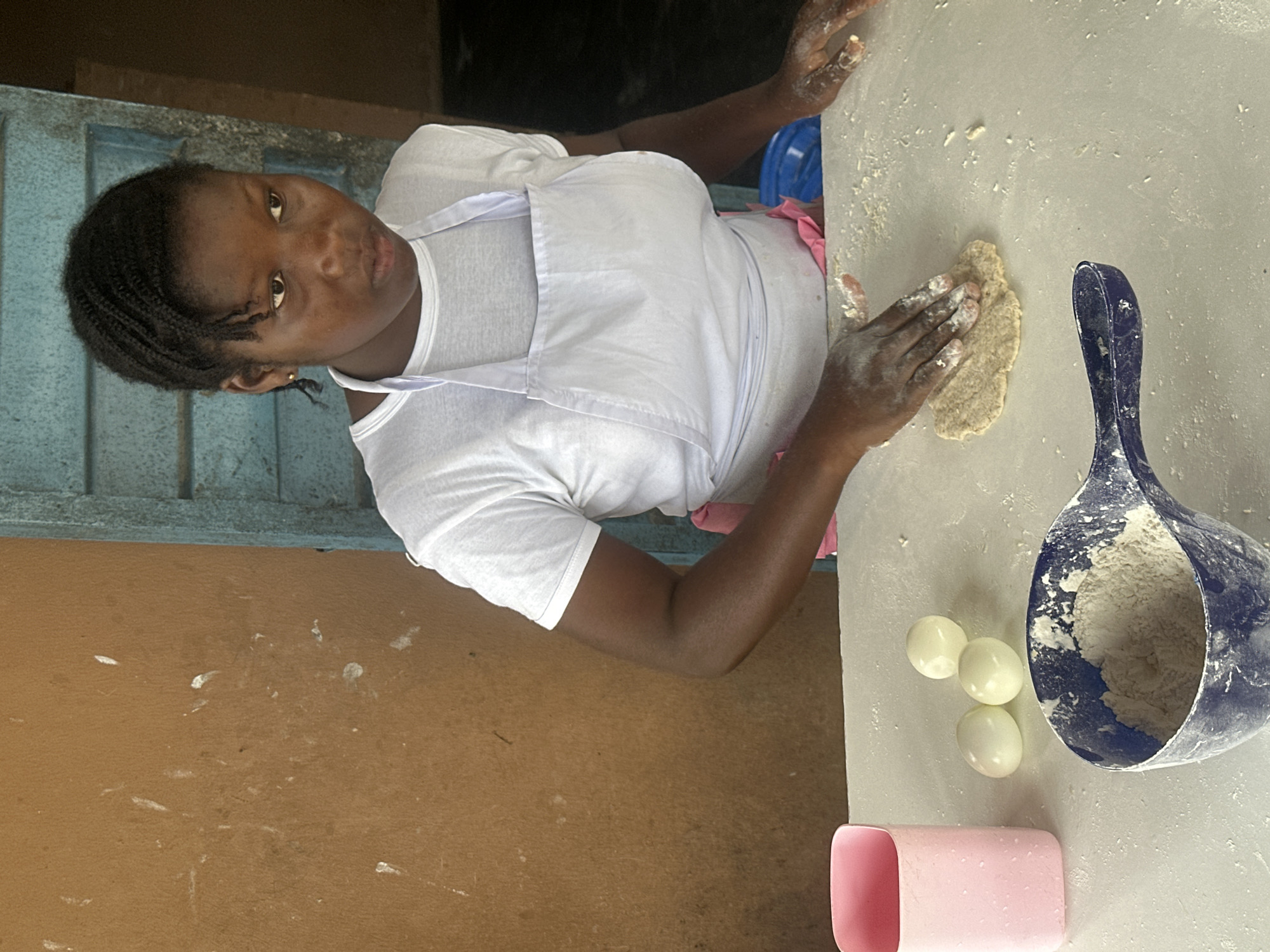 In Badagry, Nigeria, a young Black woman, with braided hair and wearing a white t-shirt and apron, kneads dough on a white table. She is focused on her task, her hands covered in flour. To her left, a pink cup and three white spherical objects, possibly eggs, sit on the table. A blue scoop, also dusted with flour, rests in the foreground. The background features a textured, brown wall and a blue, shuttered doorway. The lighting suggests an indoor setting with natural light.