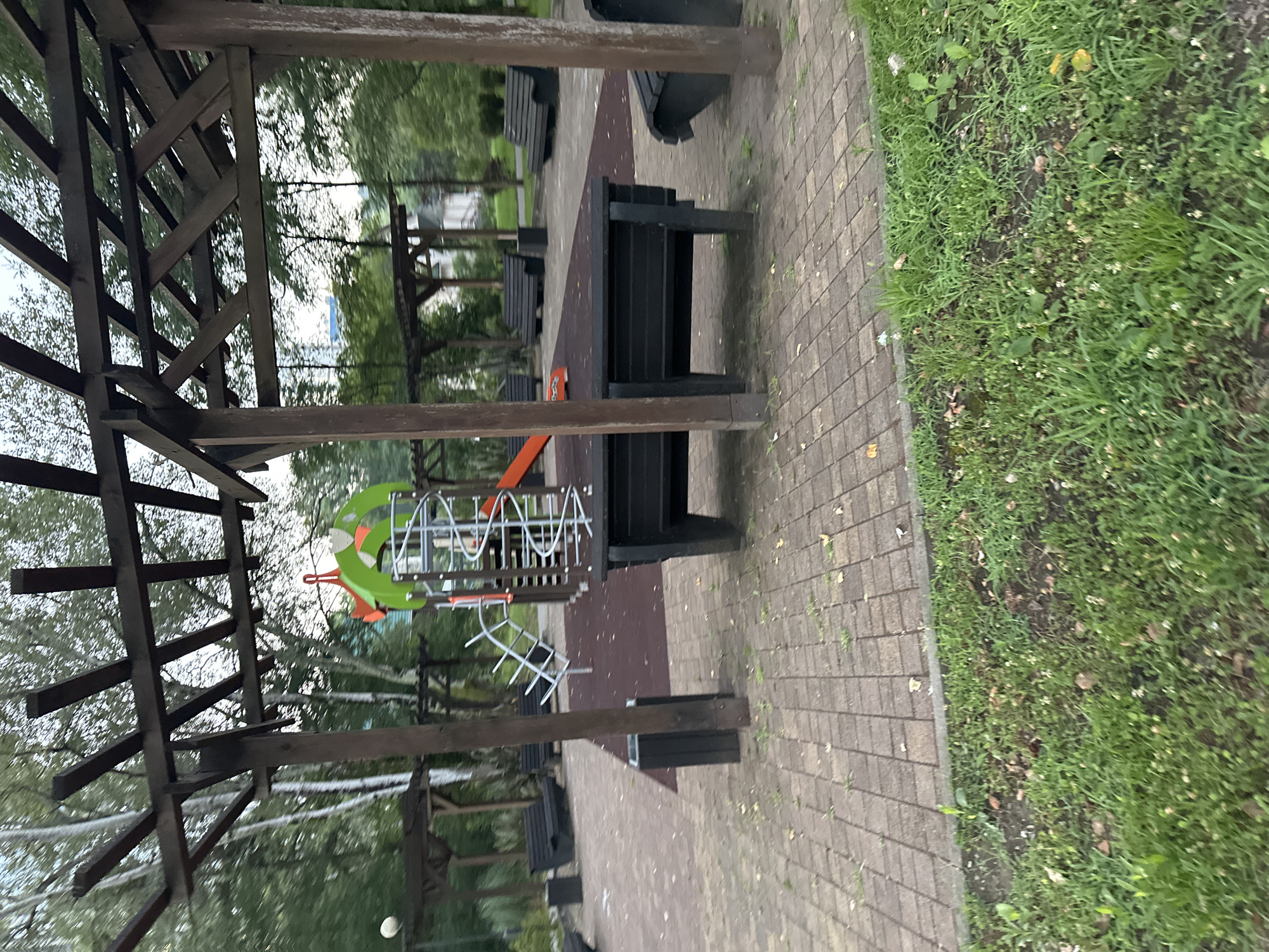 The image shows an empty playground with a wooden pergola structure in the foreground. The playground features a spiral slide, a climbing structure with metal rungs, and several dark-colored benches. The ground is covered with a reddish-brown rubberized surface, surrounded by a paved walkway made of interlocking bricks. Beyond the playground, trees and foliage are visible, with some buildings in the background. The scene appears to be set in a park or public recreational area. There are no people or animals present in the image. The lighting suggests it is either early morning or late afternoon.