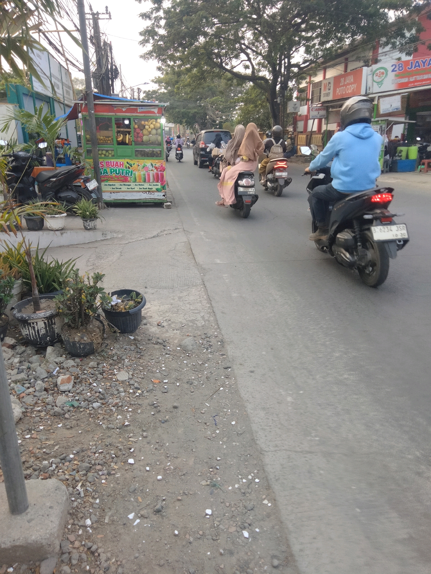 The image captures a street scene in Sepatan, Indonesia, likely during the day given the lighting. In the foreground, a grey pole stands on a concrete base, with gravel and debris scattered around it. To the left, several potted plants are visible, some appearing neglected.
The central focus of the image is a food cart selling fruit juices. The cart, painted green and red, displays various fruits such as bananas, dragon fruit, and melons. Text on the cart lists juice flavors like "Jus Alpukat," "Jus Mangga," "Jus Melon," and "Jus Jambu." Several motorcycles are parked near the cart.
Further down the street, a procession of motorcycles is moving away from the camera. The lead motorcycle has a rider in a blue hoodie and dark pants, wearing a helmet. Behind this, a motorcycle carries two passengers wearing hijabs, one in pink and the other in beige. Another motorcycle is visible further back.
In the background, buildings line the street, one with signage that includes "28 JAYA POTO COPY." Trees with dense foliage overhang the street. The road surface is asphalt, showing signs of wear and tear. The overall impression is a bustling urban environment with commercial activity and everyday traffic.