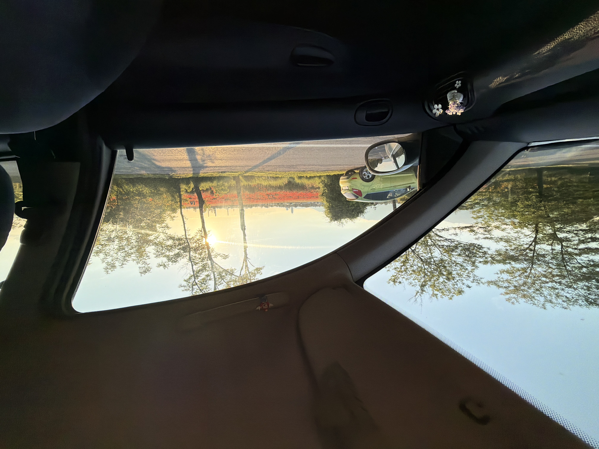The photograph is taken from inside a car, looking out through the front passenger window and the windshield. The viewpoint is low, capturing the interior of the car including the door panel with a small decorative item featuring flowers clipped to the air vent. Outside the car, a bright sun is setting or rising, casting a warm glow. A field of red poppies is visible in the distance, stretching towards a hazy horizon with a few buildings. A light green car is parked on the side of a road. Trees line the road, their leaves illuminated by the sunlight. The sky is clear with a gradient of blue and orange from the sunset/sunrise. The location is identified as Kazichene, Bulgaria. The scene depicts a scenic view from a car, likely during a road trip.