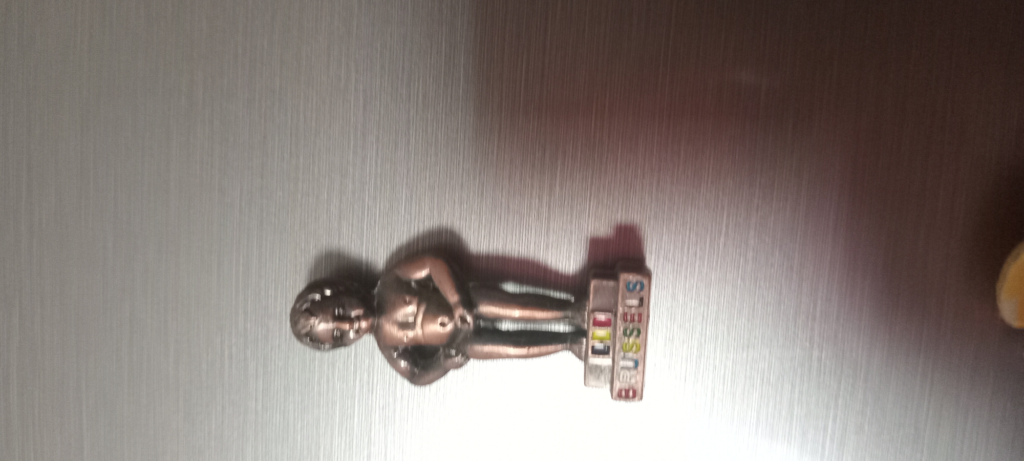 The media file is a close-up, high-angle shot of a small, bronze figurine of the Manneken Pis statue from Brussels. The figurine is placed on a brushed metal surface, possibly a refrigerator door, given the reflective quality of the surface and the presence of a shadow. The figurine is positioned horizontally, with its head pointing to the left.
The Manneken Pis figurine is detailed, showing the figure in its characteristic pose, with its left leg slightly forward. Attached to the base of the figurine are two stacked blocks of text. The top block has the letters "ILL" in black on a white background, and the bottom block has "BRUSSELS" in colorful letters (blue, yellow, red, green) on a white background. The lighting suggests an indoor environment, possibly with artificial light casting a shadow to the right of the figurine. The overall impression is that of a souvenir item.