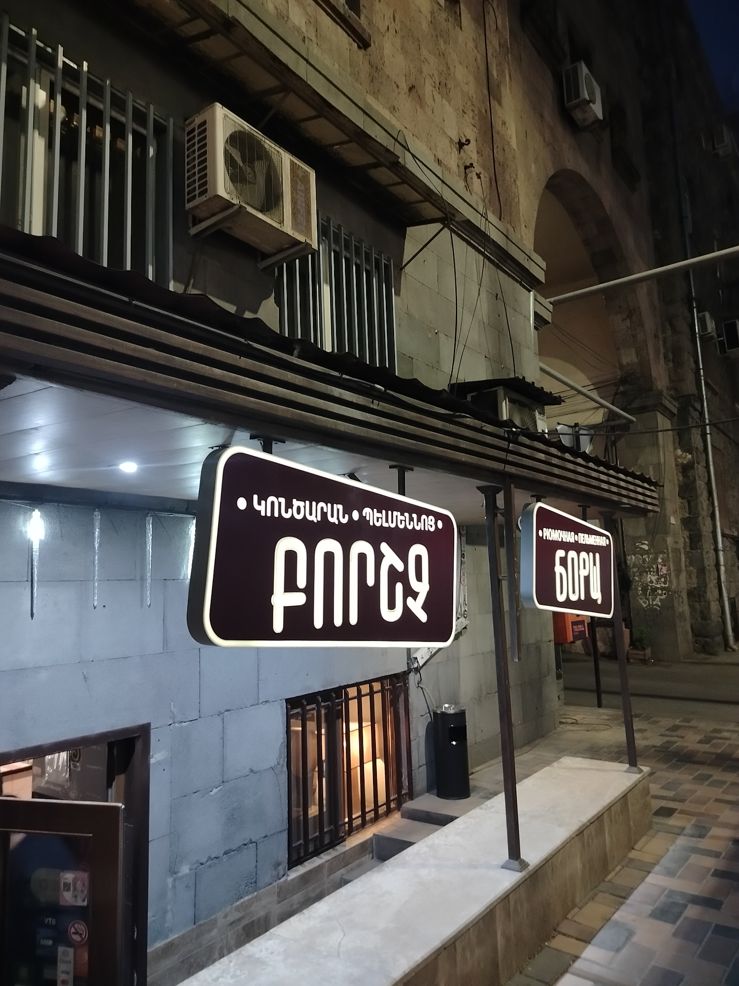 This appears to be an outdoor shot of a business in Yerevan, Armenia, at night. The business has two signs in front, both written in a Cyrillic script and in Armenian.
The sign to the left appears to read “ԿՈՆՏԱՐԻ ԴԵՏԵԿՏԻՎ ՊՈՐՏ” and the sign on the right appears to read “РОМОЧНАЯ ПЕЛЬМЕННАЯ БОРЩ”.
Behind the signs is a gray building with bars on the windows. There are two air conditioning units mounted on the exterior wall. A black trash can sits on a step between the two signs.
The pavement in front of the business is made of mixed stone and concrete. The business appears to be located on a street in Yerevan.