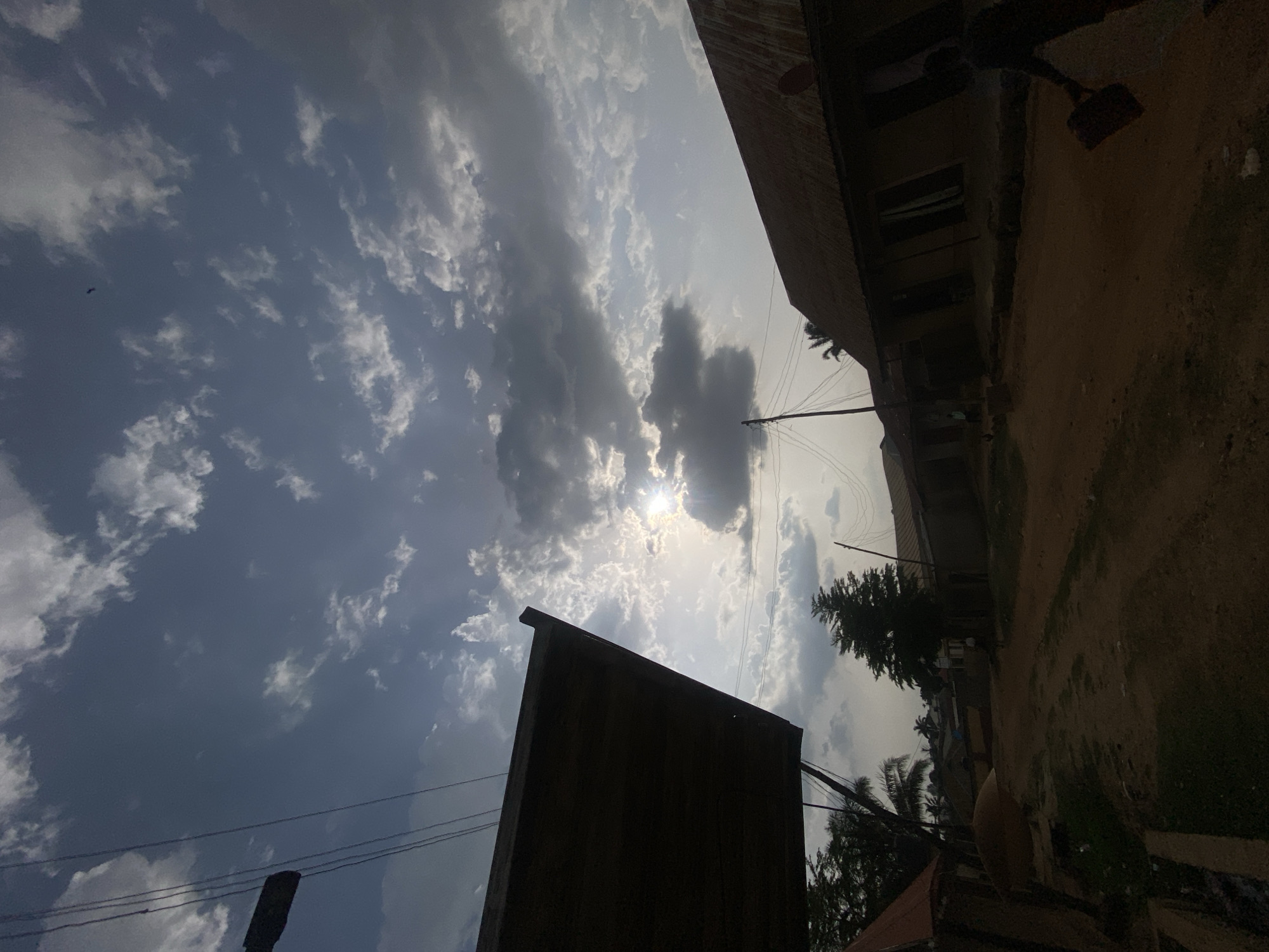 The photograph, taken in Narayi, Nigeria, depicts a partially overcast sky with the sun shining through a break in the clouds. A dark silhouette of a building occupies the foreground. In the background, a building with windows and a corrugated metal roof is visible along with a dirt yard. Power lines stretch across the sky. Trees are scattered throughout the scene.