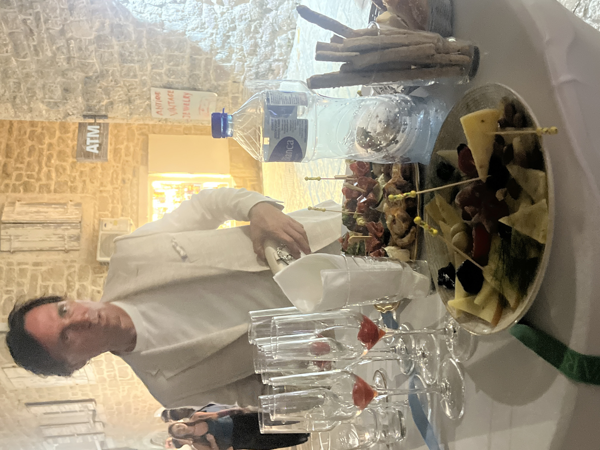 A man in a white suit and t-shirt is standing behind a table laden with appetizers and drinks. The table is set with champagne flutes, some containing red liquid, a platter of cheese and olives, and a dish with small bites. A bottle of water and breadsticks are also visible. In the background, a sign for an ATM and another sign that reads "ANTIQUE VINTAGE JEWELRY" are mounted on a stone wall, suggesting the location is a street in the old town of Budva, Montenegro. The man appears to be pouring from a bottle, possibly champagne, into one of the flutes. Two women are partially visible in the background, walking away from the camera. The lighting suggests it is evening.