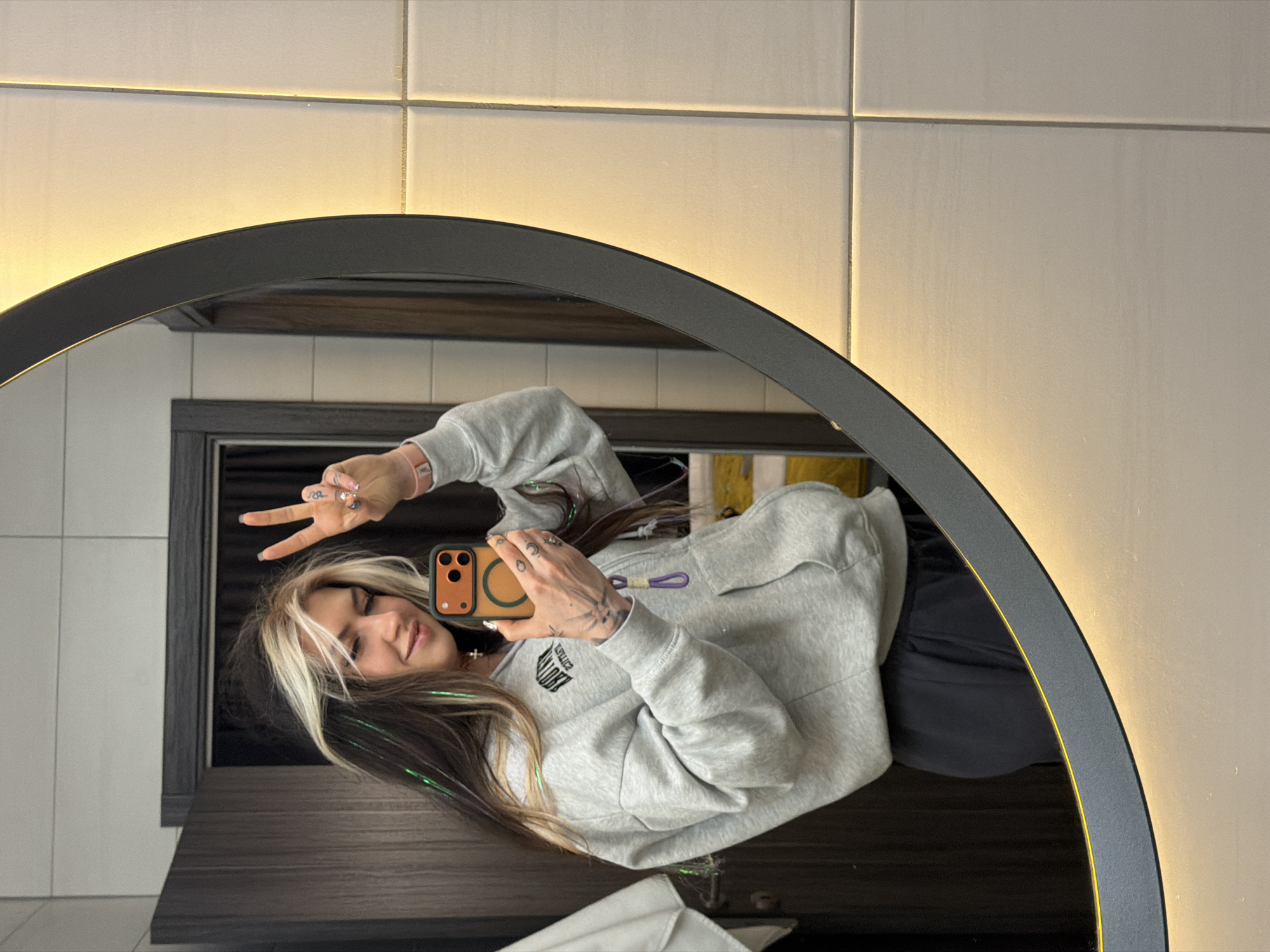 A person is visible in a mirror in what appears to be a bathroom setting. The person, with white highlights in their brown hair, is wearing a gray sweatshirt and holding a mobile phone with an orange case, presumably taking a selfie. They are making a peace sign with their fingers. The mirror has a dark gray circular frame. The background consists of off-white rectangular tiles and a dark gray shelf. The location of the image cannot be reliably determined as Palilula Urban Municipality, Serbia, based on the available visual information.