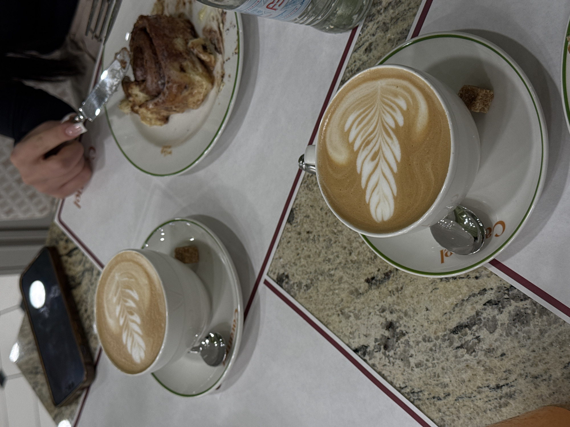 The image captures a scene at a cafe in Yerevan, Armenia. The setting is a table with a marble countertop, partially covered by a white placemat bordered with a thin red line. On the placemat, there are two cups of coffee, each with latte art featuring a leaf pattern. One cup is positioned closer to the camera and has a brown sugar cube on its saucer. A spoon is visible on each saucer.
In the upper left corner of the image, a partially eaten pastry sits on a plate. A hand holds a knife, seemingly about to cut into the pastry. A water bottle is situated next to the plate. In the lower left corner, a smartphone is present.
The scene suggests a casual dining experience, likely a coffee break. No specific interactions between people are evident in the frame.
