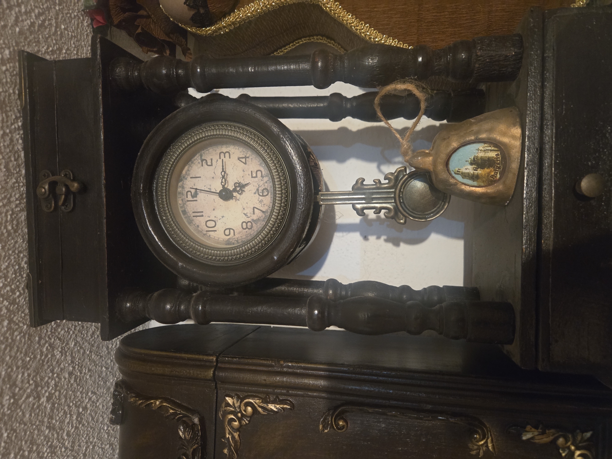 An antique clock sits at an angle on a decorative dark wooden surface. The clock face is circular with Roman numerals and the hands are pointing to approximately 1:30. A decorative bell with a picture of a cityscape, labeled "Bulgaria," hangs next to the clock. The wood is carved with a golden accent. The location is unknown, and there are no people present in the image.