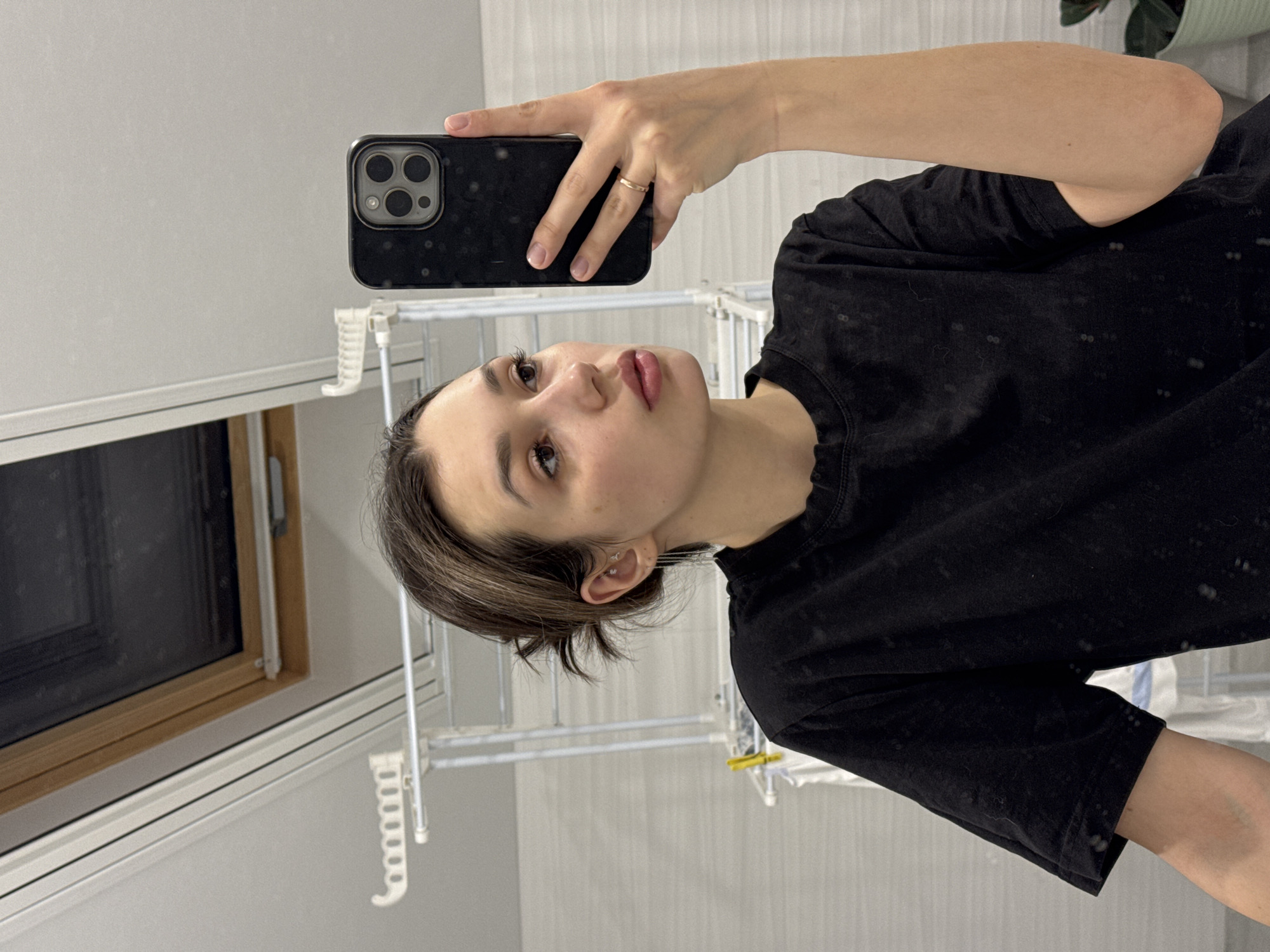 A young person is taking a selfie in what appears to be an indoor environment, possibly a residence. The person is holding a black smartphone with a multi-lens camera setup. They are wearing a black t-shirt. The background includes a window with a wooden frame, a drying rack, and a partial view of a green plant. The city and specific location within it are unknown.