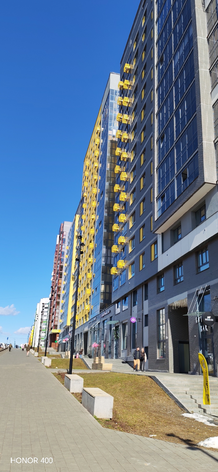 The image shows a street scene in Izhevsk, Russia, featuring a row of modern apartment buildings with colorful facades. The buildings vary in height and architectural style. One building has yellow balconies and accents, while another is predominantly gray with blue-tinted windows.
A paved pedestrian walkway runs along the base of the buildings, with benches and small landscaped areas separating it from the building entrances. Several people are walking on the sidewalk.
A business is visible in the building's ground floor. The sky is clear and blue. The overall scene suggests a modern urban residential area.