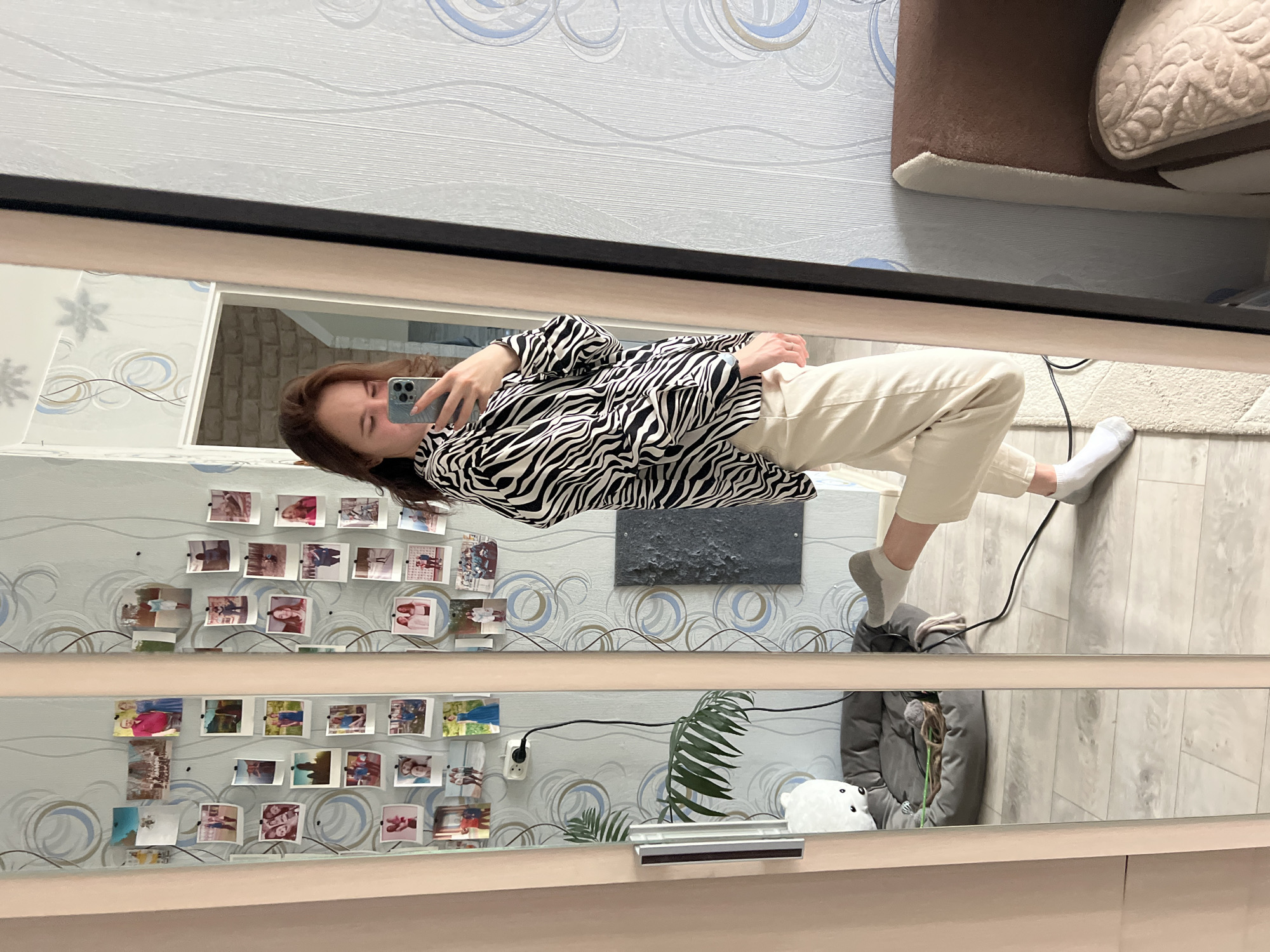 A full-body mirror selfie captures a young woman standing indoors, reflected in a large, light-framed mirror. The woman has brown hair and is engrossed in her smartphone, which she holds up to take the picture, with her gaze directed towards the screen. She is dressed in a striking black and white zebra-print long-sleeved shirt, light beige trousers, and white socks, with one grey slipper visible on her right foot.
The background, visible in the mirror's reflection, reveals a wall covered with patterned wallpaper featuring light blue, white, and grey swirls, alongside subtle floral designs. Numerous small, printed photographs, resembling a collage of personal memories, are affixed to the wall. Above this photo display, a rectangular piece of dark grey, textured wall art is mounted.
The floor appears to be light-colored wood, partially covered by a white, fluffy rug. Various items are visible around the room's lower sections: a white electrical outlet with a black cord plugged into it, a green leafy plant, a white plush toy shaped like a polar bear, and a grey padded jacket or coat resting on the floor. Part of a brown, upholstered furniture piece (possibly a sofa or armchair) is also visible in the upper left of the reflection. The scene portrays a casual and personal living space, likely a bedroom or living room, located in Chaikovsky, Russia. There is no visible text that can be clearly read, and the time of day is not discernible.