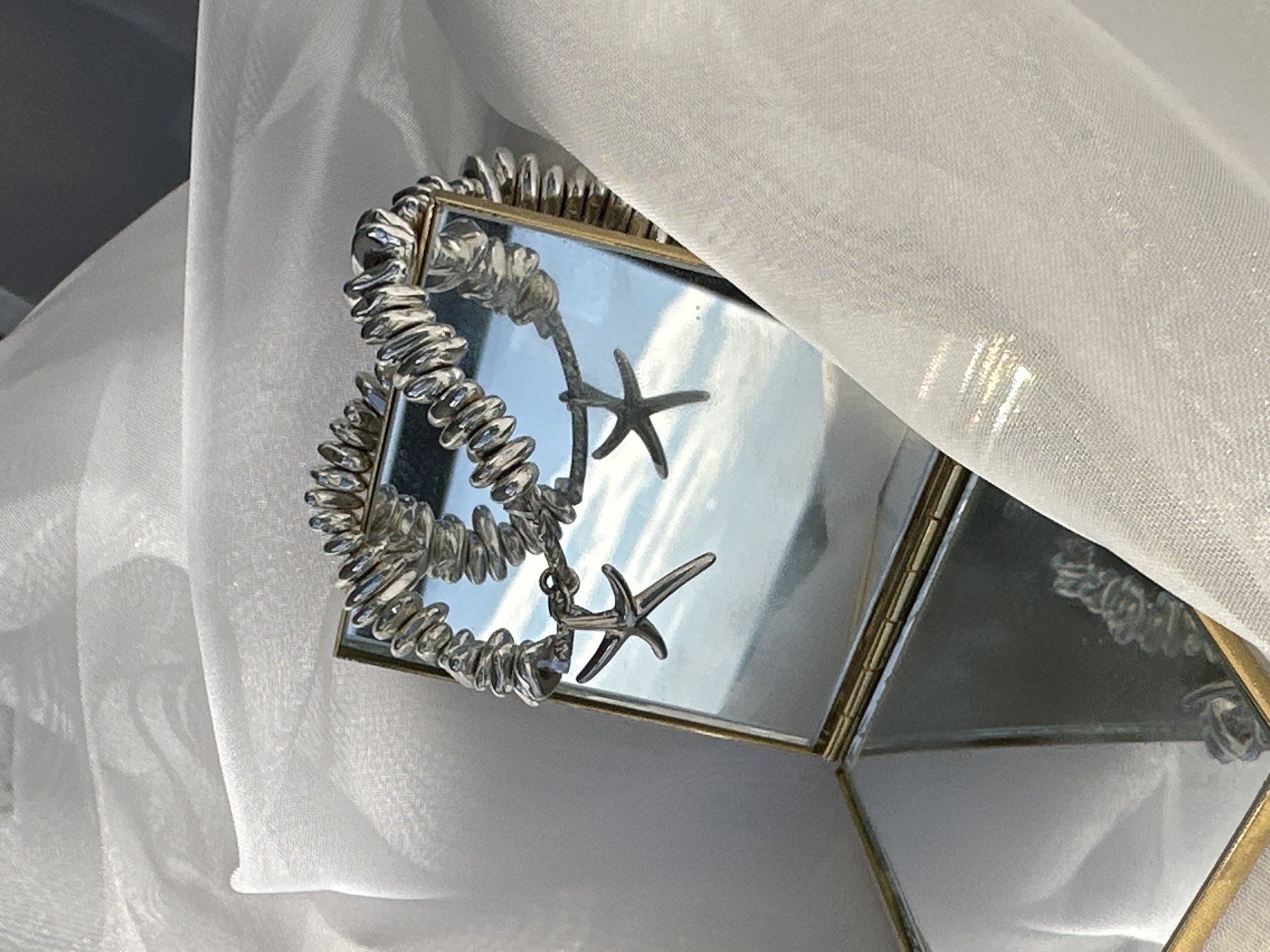 A close-up captures a silver bracelet with starfish charms draped over a small, multi-paneled mirrored box with a gold trim. The bracelet is composed of irregular silver beads strung together. The box reflects a bright, overcast sky. White, sheer fabric is draped in the background. The location is Unknown, Unknown.