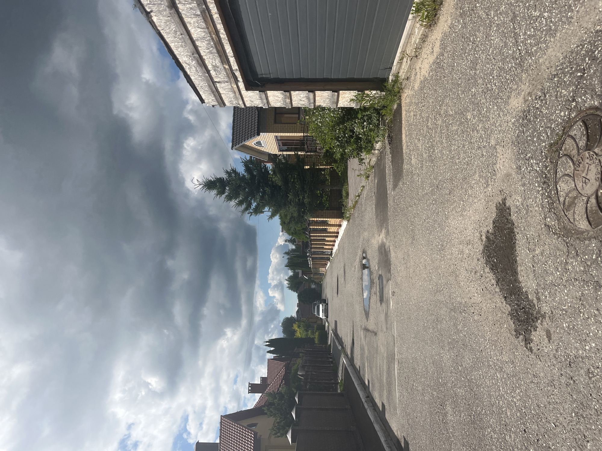 The image depicts a street scene in Orekhovka, Russia, with a cloudy sky overhead. The asphalt road is wet in patches, suggesting recent rainfall. On the right side of the road, there is a large garage with a grey sectional door and a wooden structure above it. To the left of the garage, a residential building with a yellow facade and a dark roof is visible, with a tall evergreen tree in front of it. Further down the street, on the left side, a row of houses with red tiled roofs and brown fences line the road. A white car is parked in the middle of the street. The overall impression is that of a residential area on a possibly overcast day.
